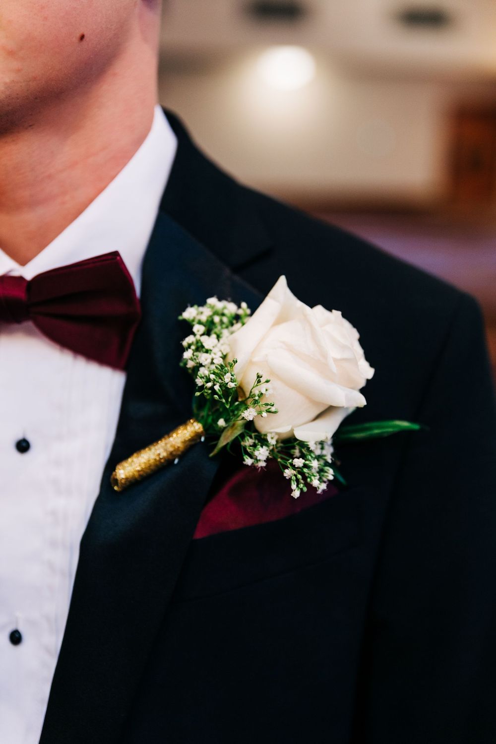 Elegant white rose and baby's breath boutonniere with burgundy bow tie on black tuxedo lapel.
