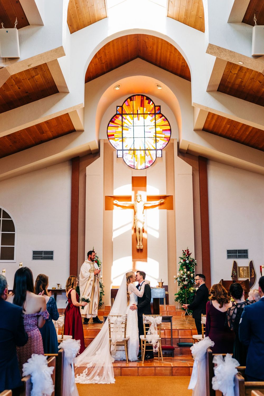 A wedding ceremony takes place in a Catholic church with a large stained glass window and wooden beams overhead.