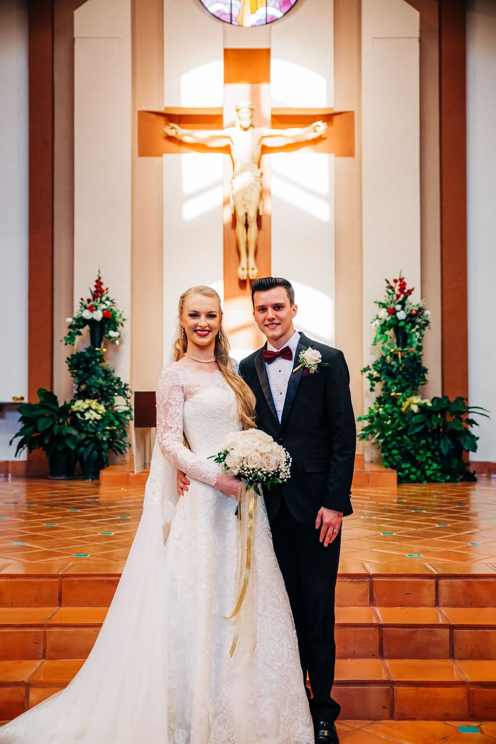 A formal wedding photo taken at the church altar with stained glass window and crucifix in background.