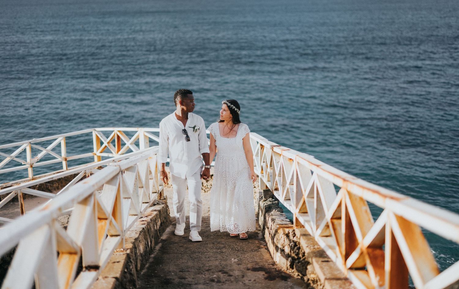 Romantic couple in white attire walking on a seaside pier during their coastal wedding.