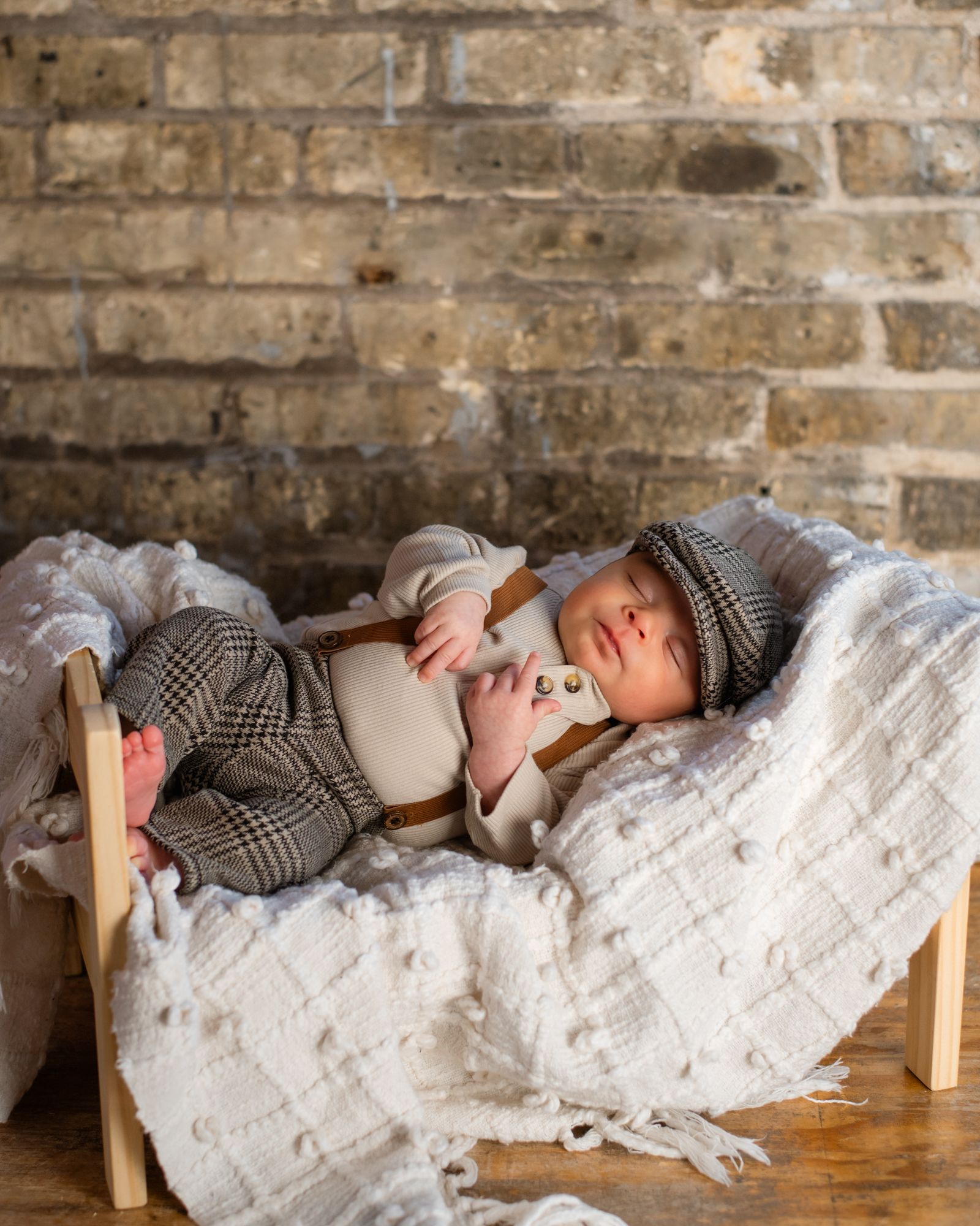 Sleeping baby in vintage outfit and cap rests on textured blanket against brick wall.