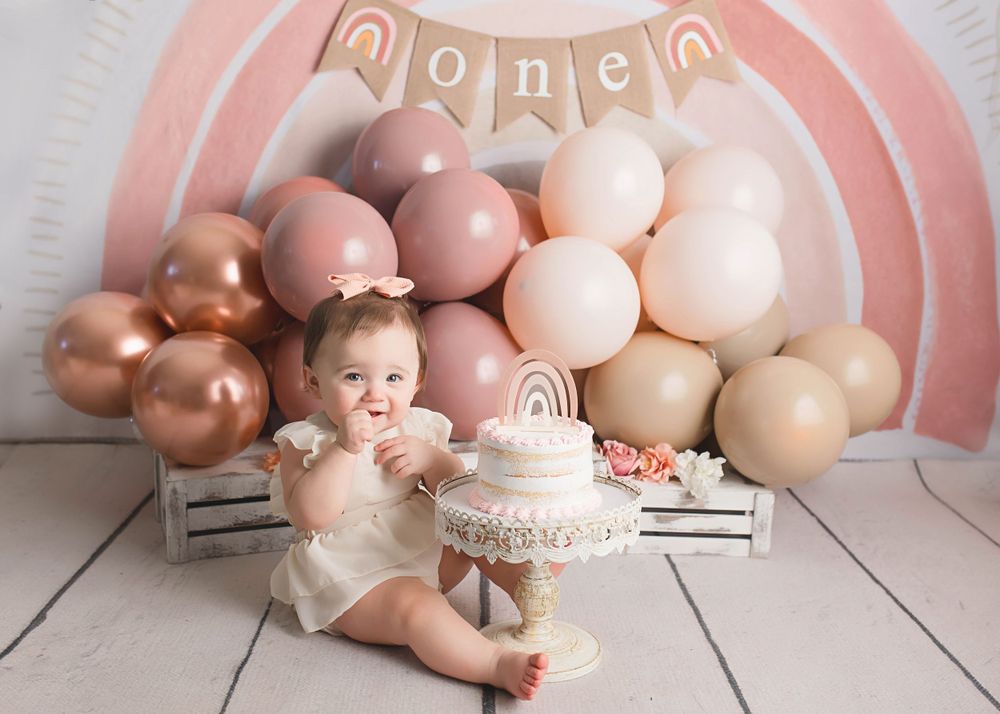 First birthday setup with ombre balloon arch, white cake, and baby in cream dress.