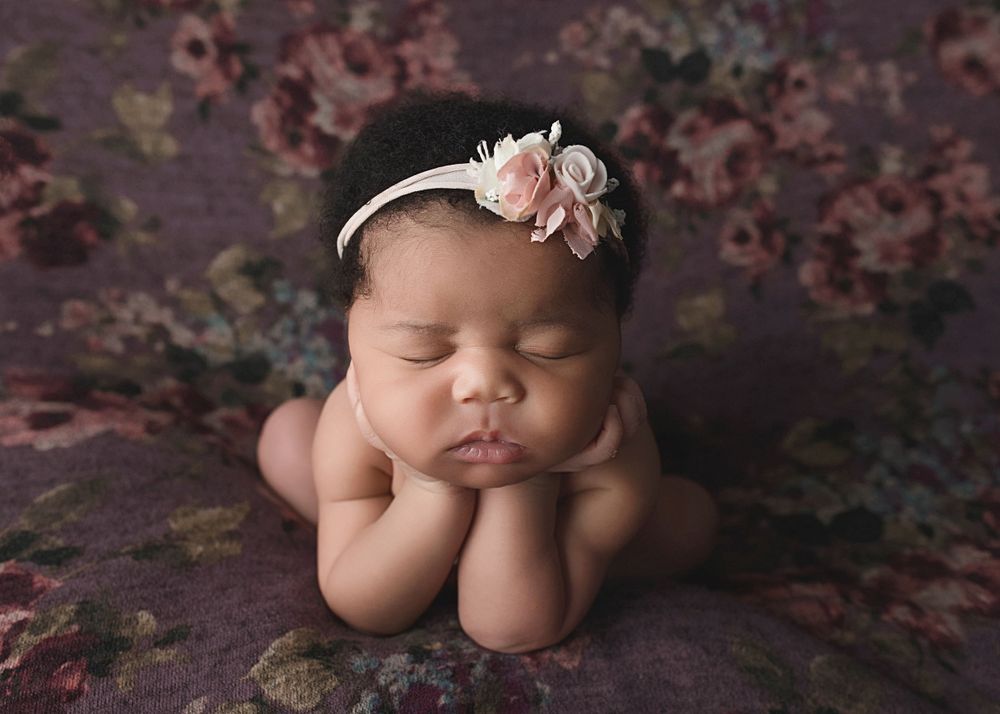Sleeping newborn baby posed with hands under chin wearing pink floral headband.