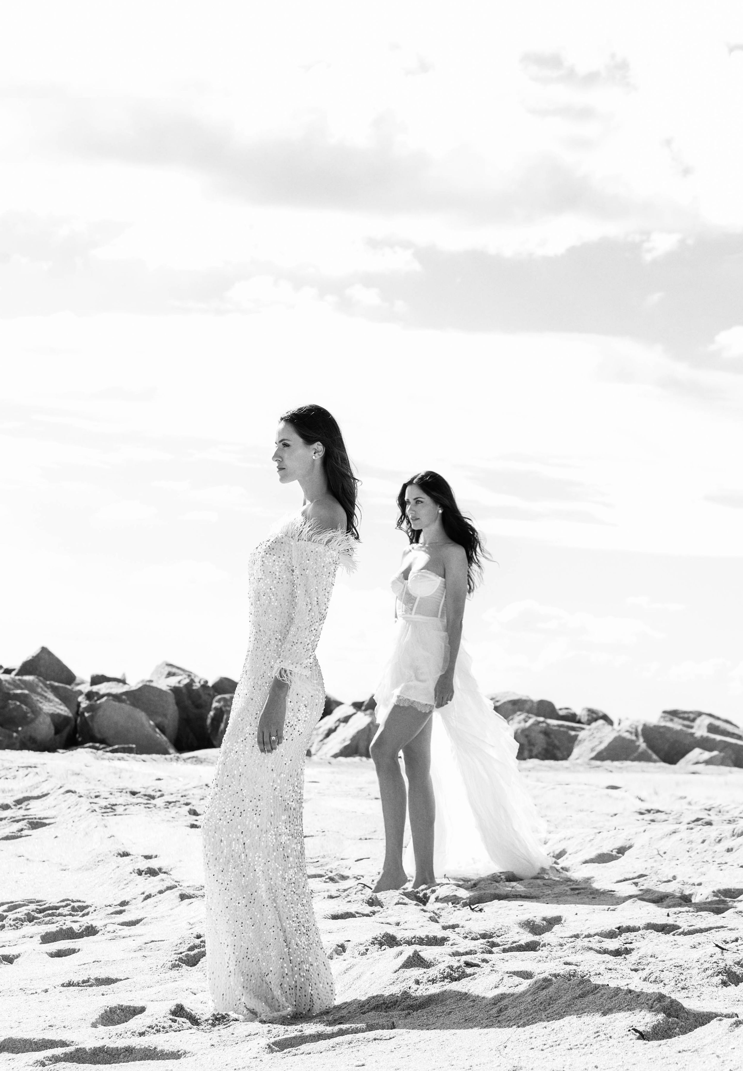 Two brides in elegant white dresses stand on a beach with rocky outcrops and cloudy sky in black and white photography.