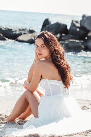 Woman in white dress poses elegantly on beach with rocky coastline and turquoise waters in background.