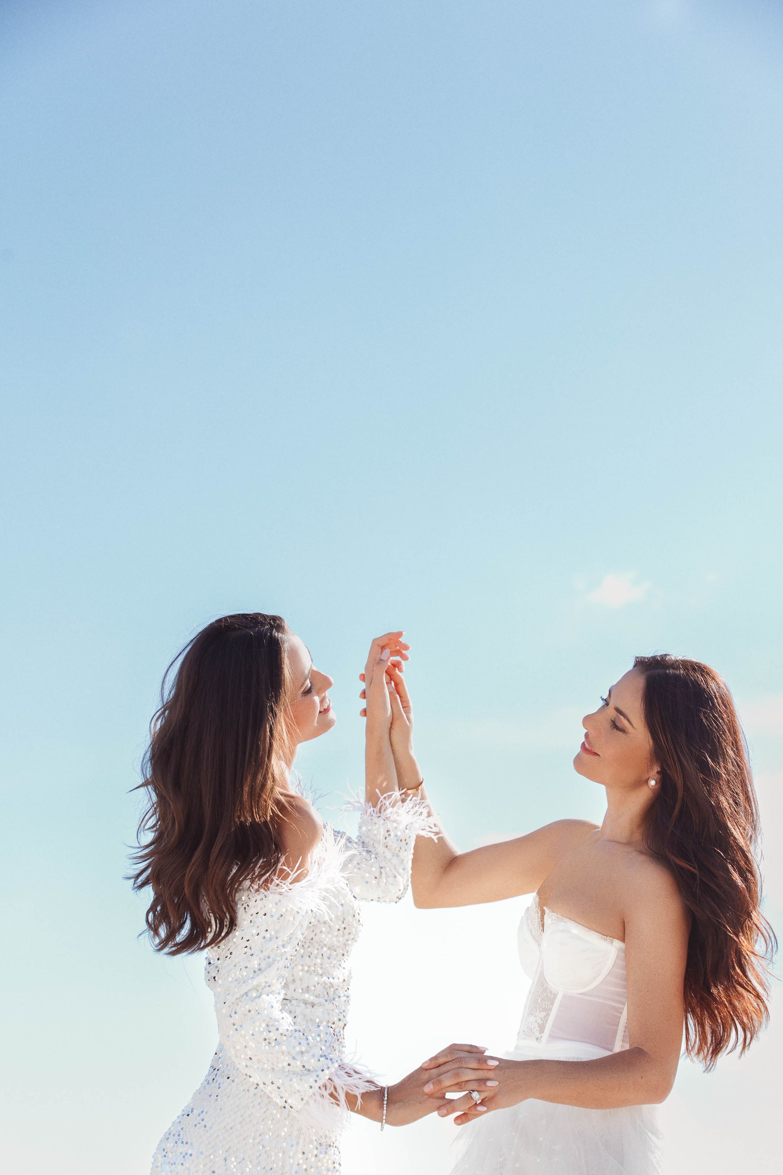 Two people in white dresses dance together joyfully against a bright blue sky background.