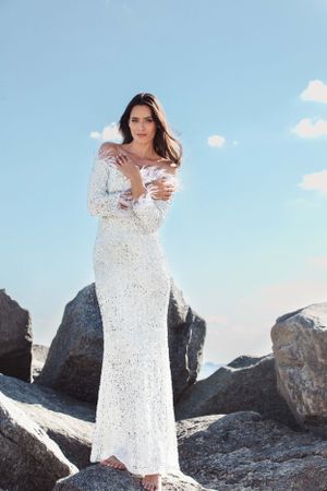 A romantic outdoor photoshoot featuring a lace white wedding dress against rocky coastal terrain and blue sky.