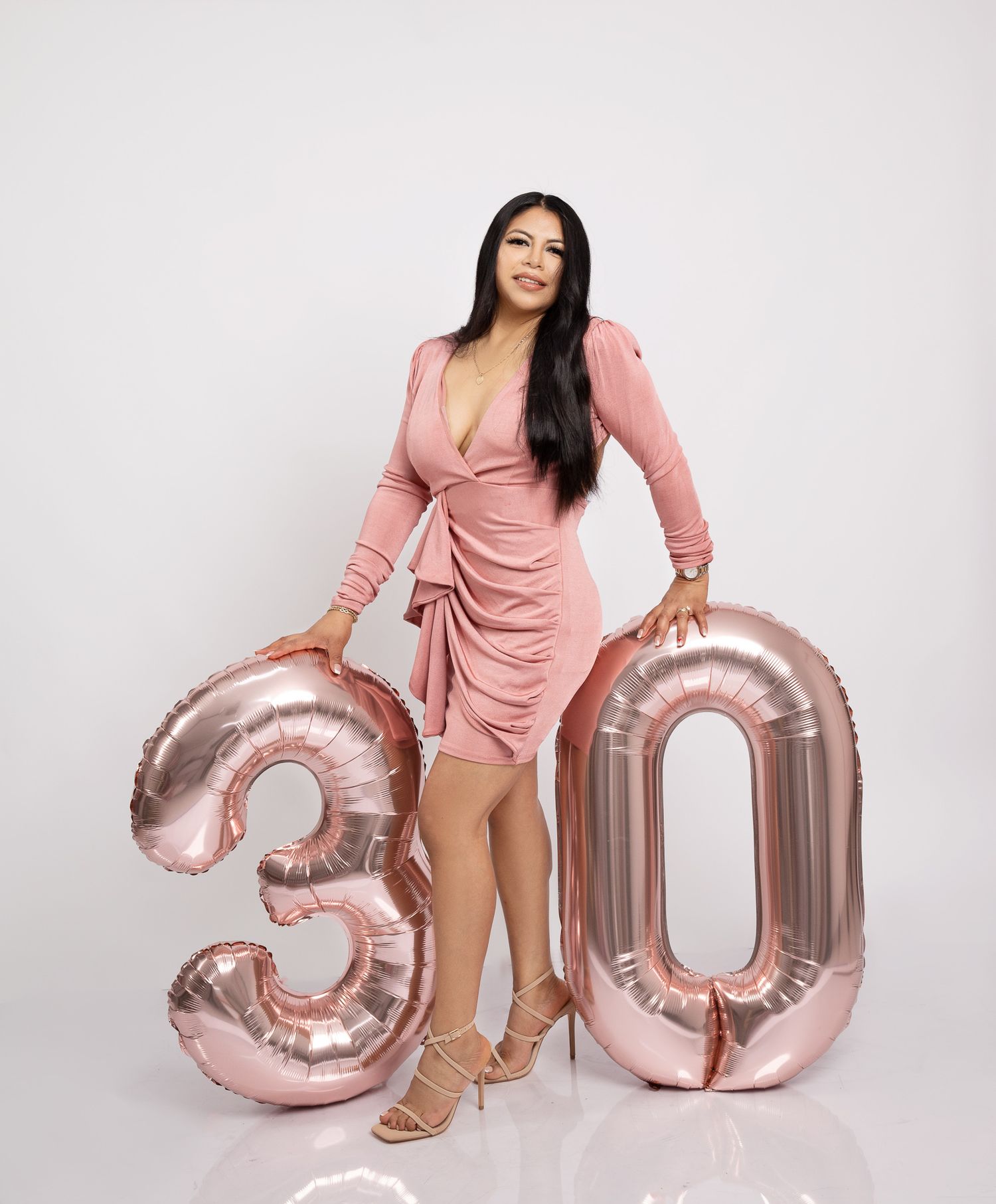 A person in a pink ruched dress posing with rose gold number 30 balloons against a white backdrop.