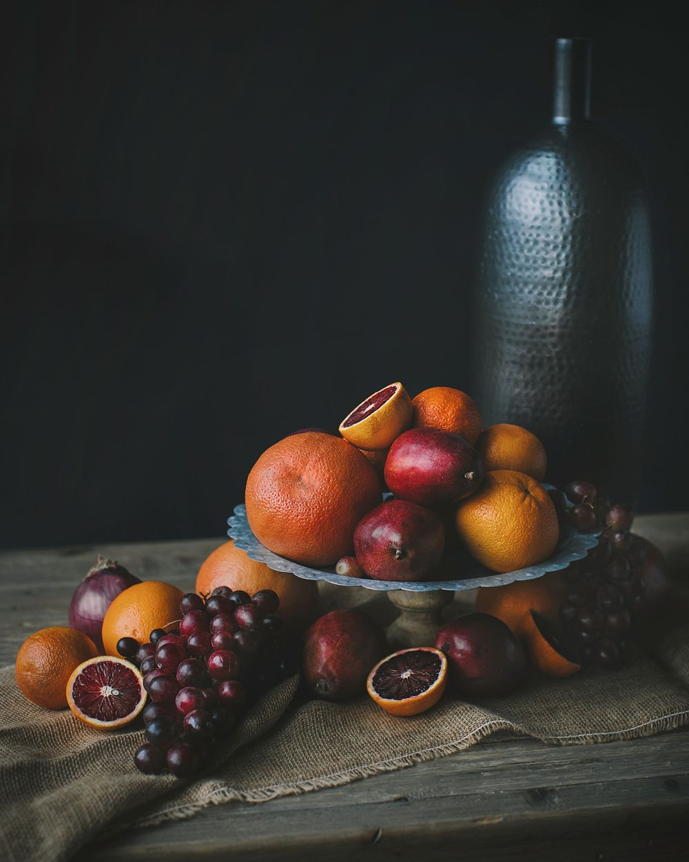 Dark moody still life of mixed citrus fruits and grapes in a bowl beside a metal vase.