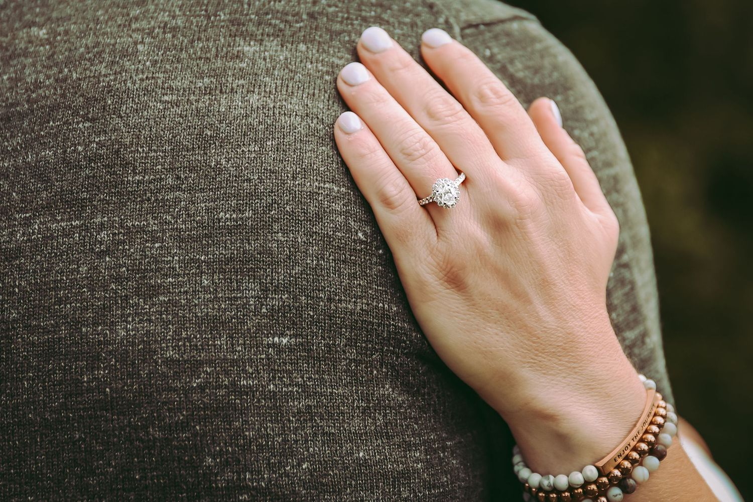 A close-up shot of a hand wearing an engagement ring and bracelet against a gray sweater.