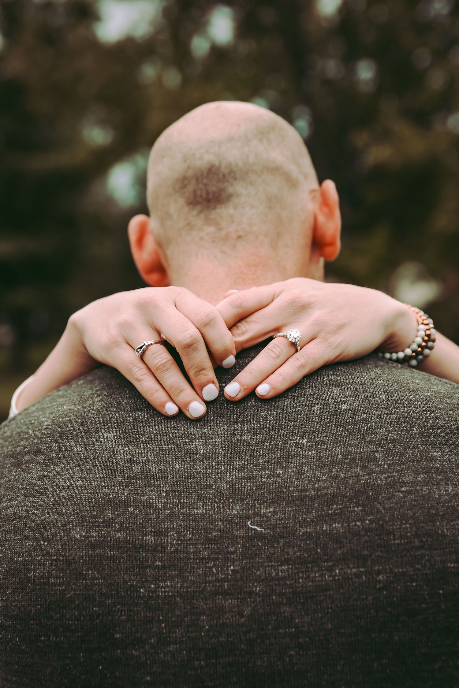 A close-up of hands wearing rings embracing around shoulders in a gray sweater.