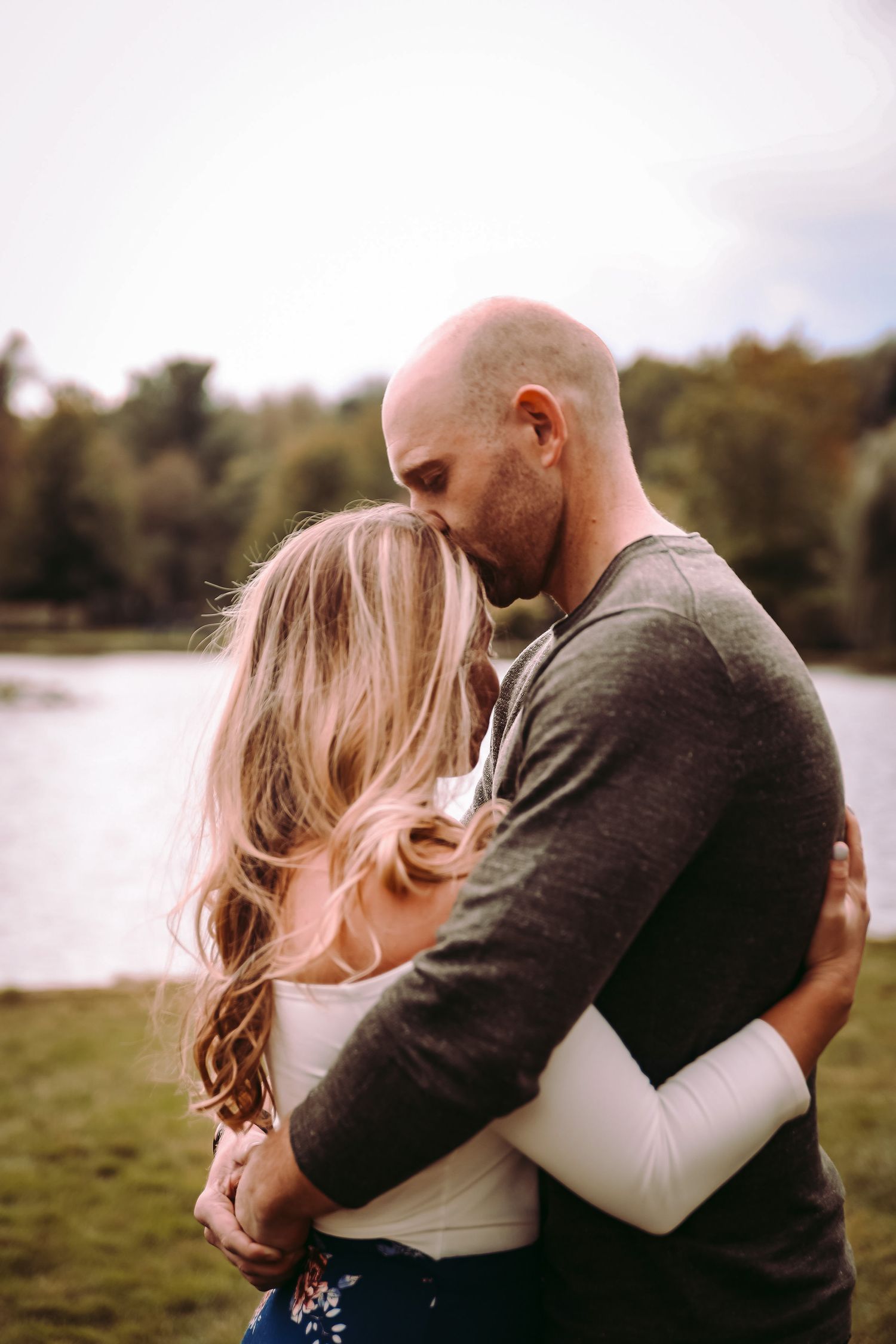 A romantic embrace between a couple beside a tranquil lake on a sunny day.