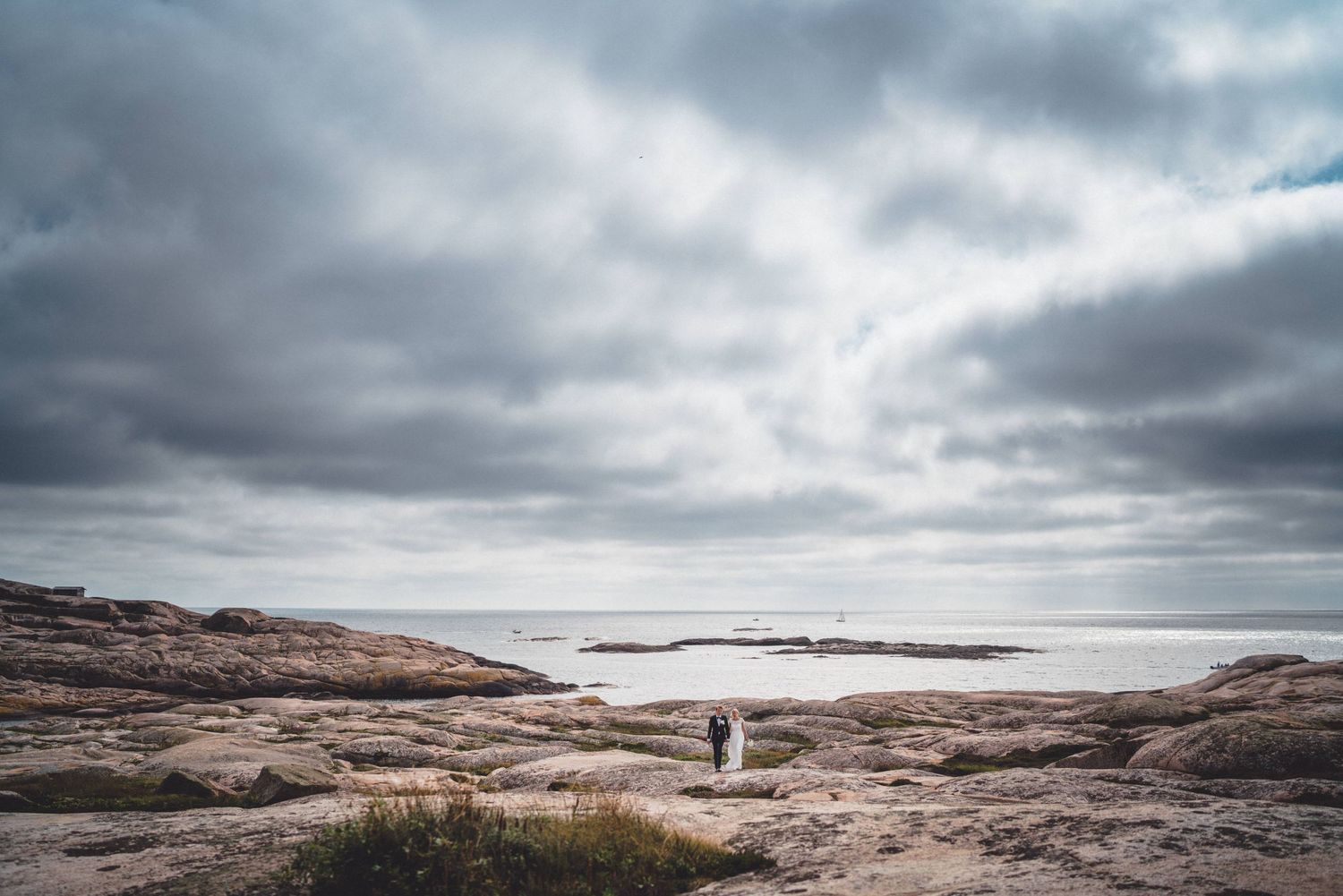 Brudpar vandrar bland bohusgraniten på Hållö utanför Smögen i Bohuslän med en segelbåt på horisonten och moln på himlen
