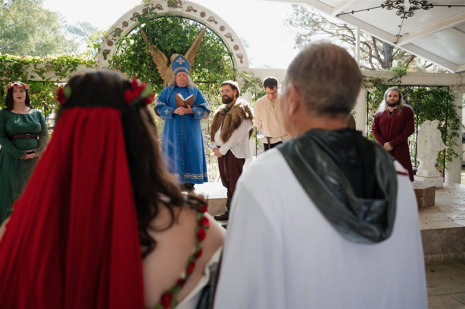 Wedding guests in historical costumes gather under decorated outdoor pavilion.