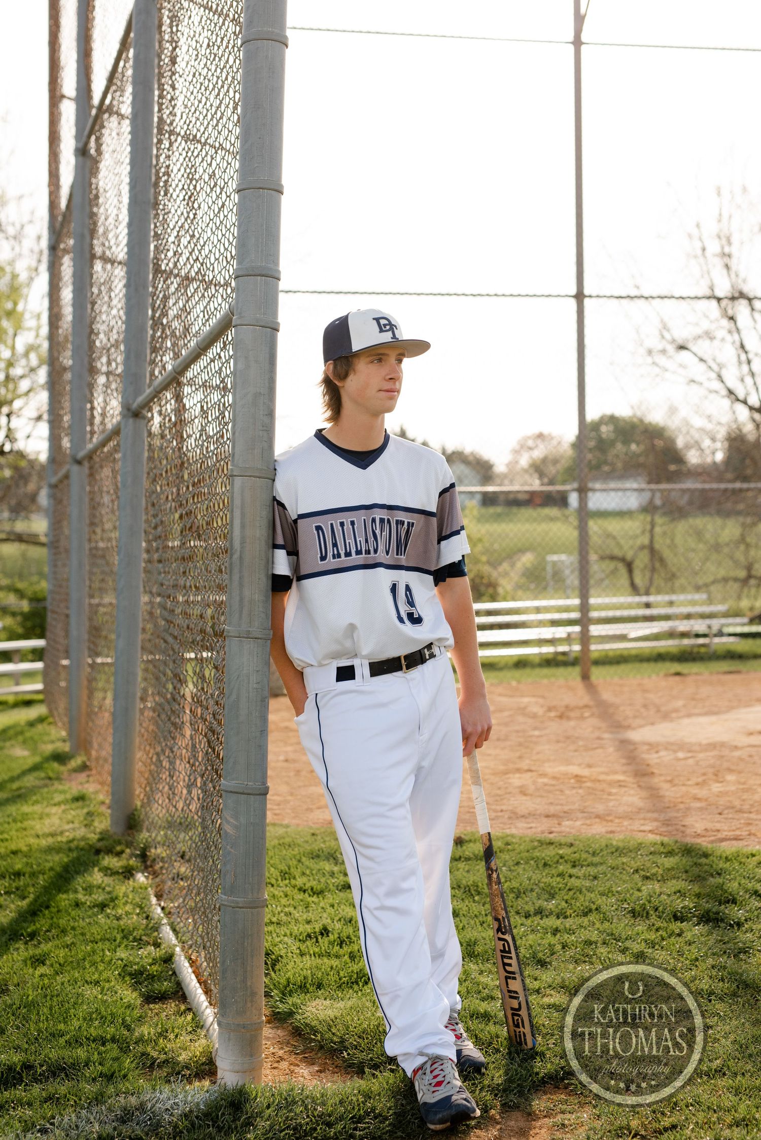 Baseball player in white uniform stands by fence at baseball field.