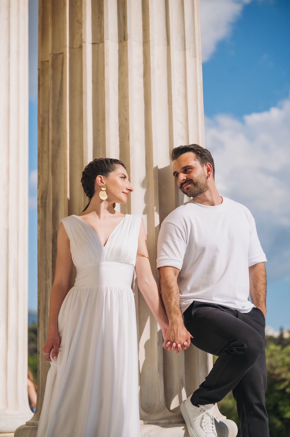Couple in white and casual attire pose romantically between classical columns on a sunny day.