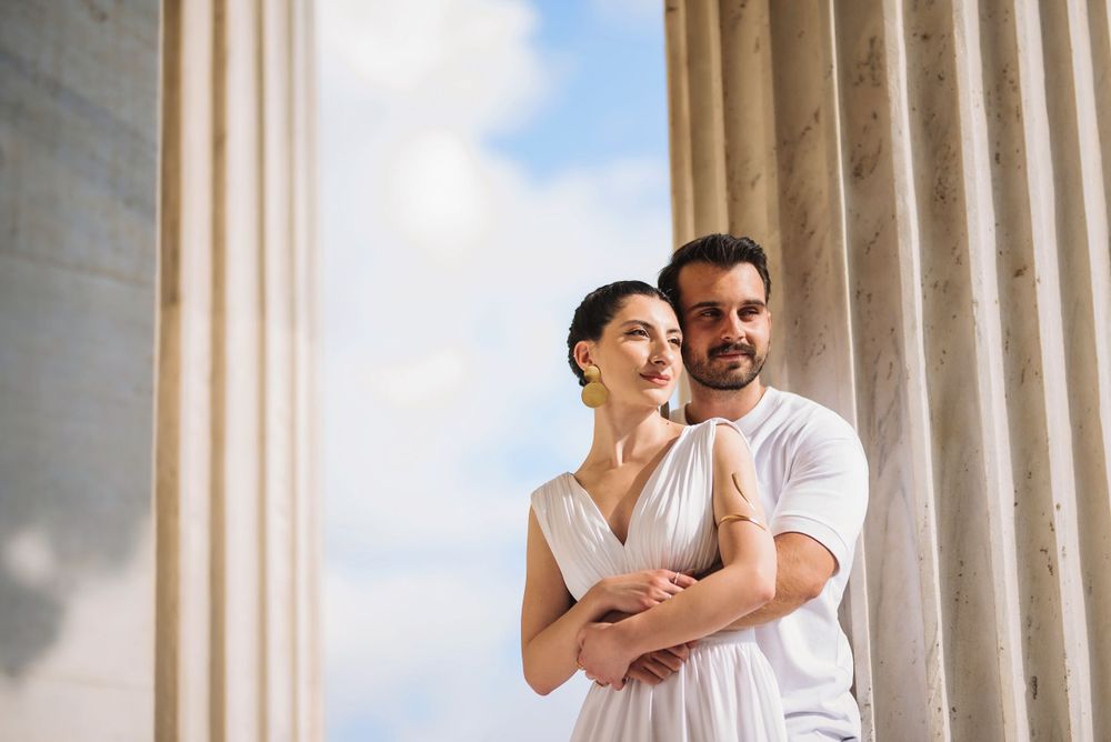 Couple in white outfits embracing against classical columns and blue sky background.