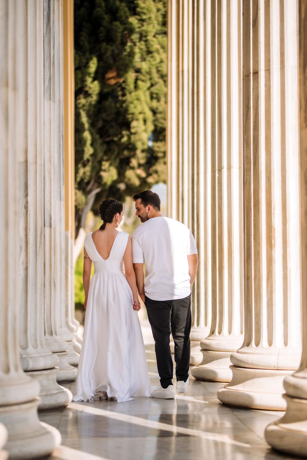 Couple in white walking between classical columns in elegant architectural setting.