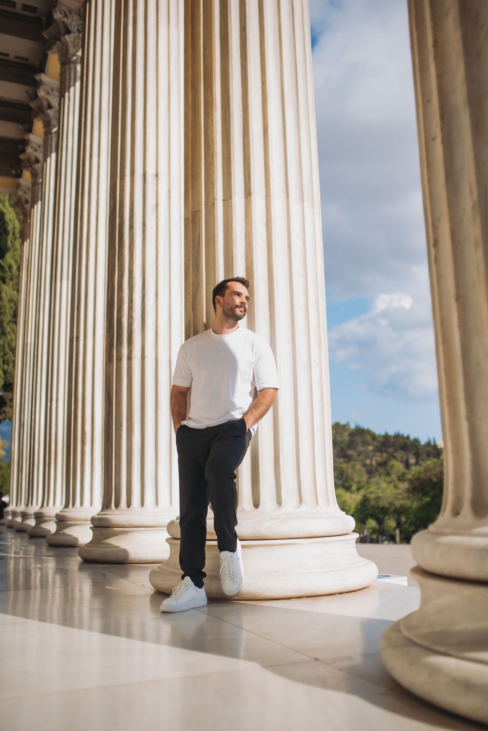 Man in white t-shirt and dark pants stands confidently between grand marble columns.