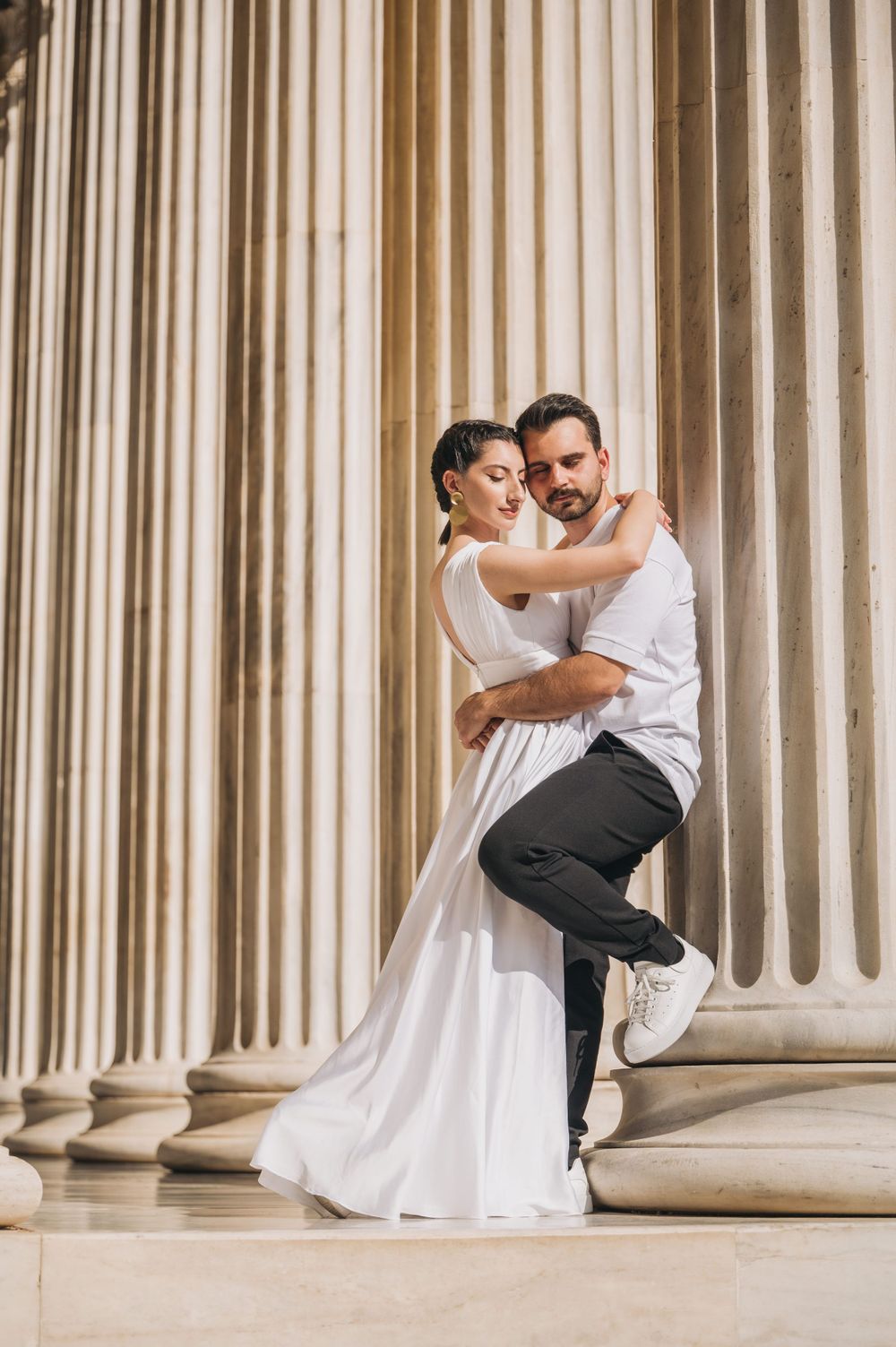 Elegant couple in white dress and casual wear posing romantically by classical columns.