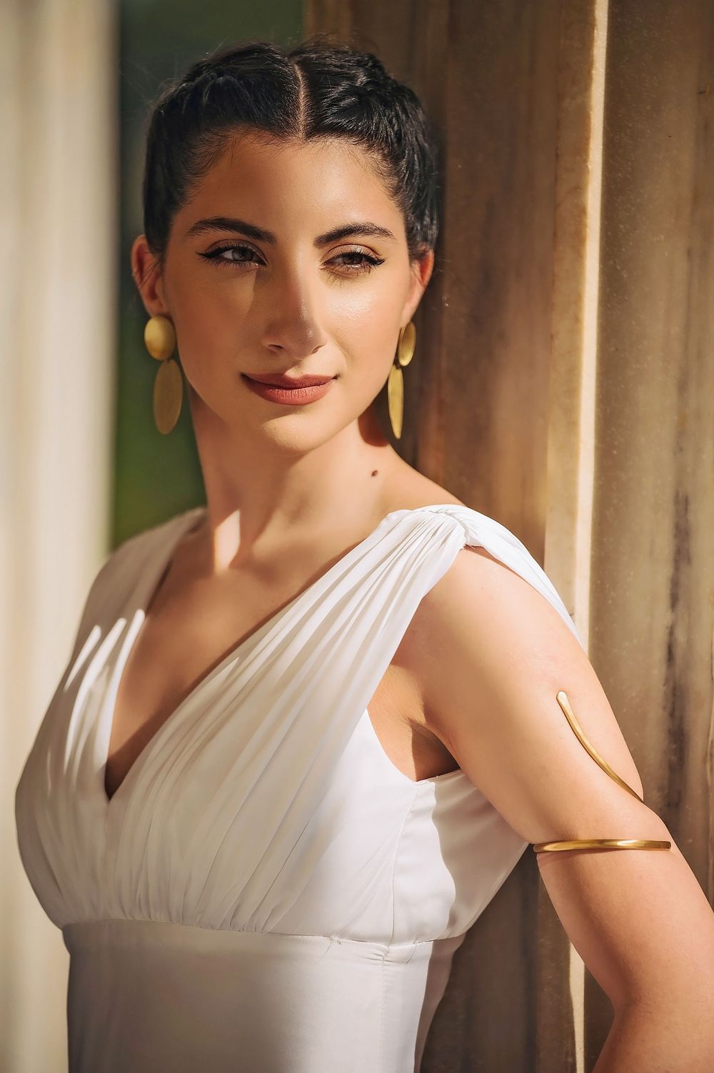 A woman in a classic white dress poses elegantly against a columned backdrop with gold earrings.