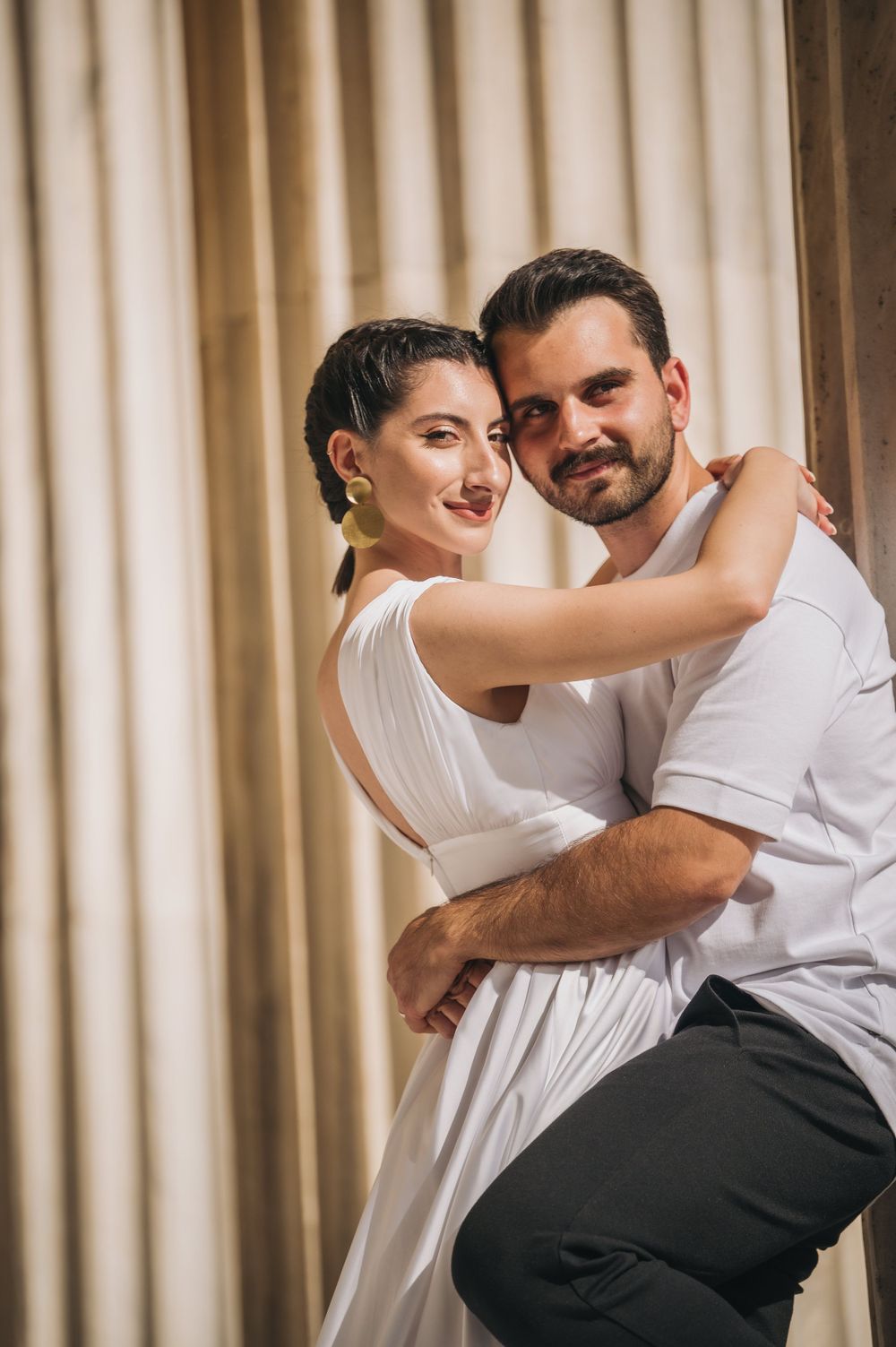 Couple sharing a tender embrace in white and casual attire against architectural backdrop.