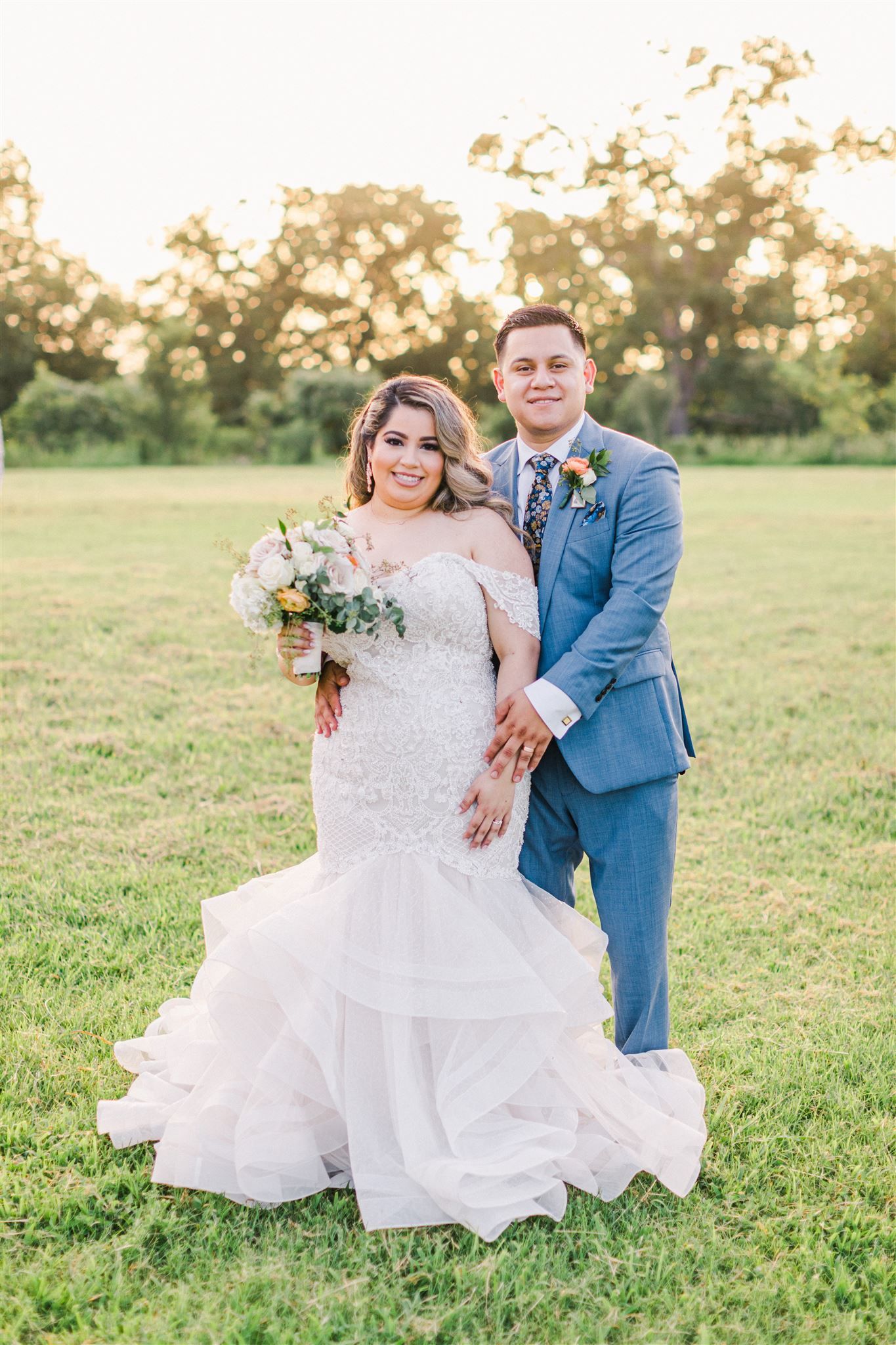 A wedding couple poses in a sunlit field with the bride wearing a lace mermaid gown and groom in a blue suit.