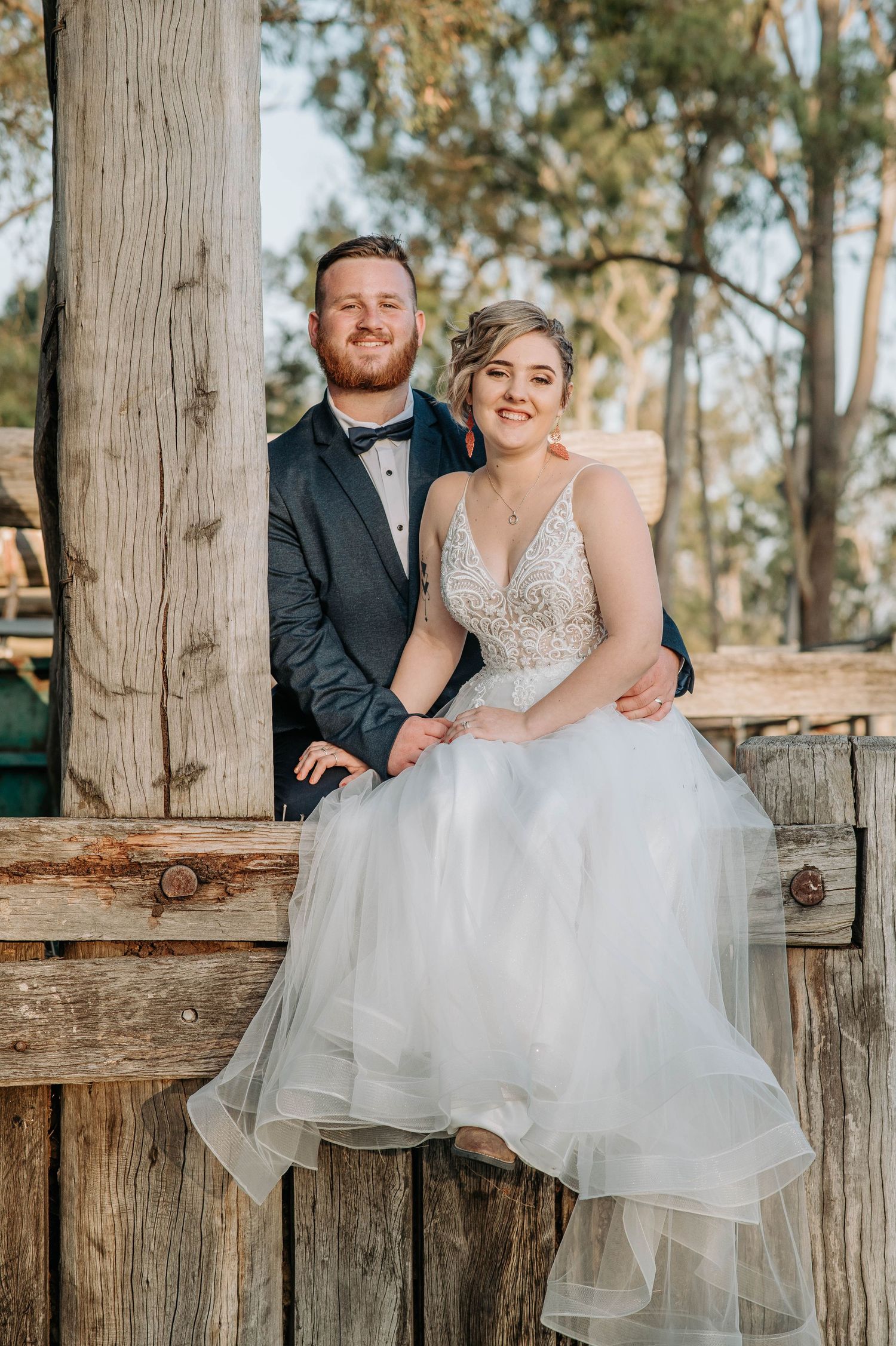 Wedding couple seated on rustic wooden porch railing in natural setting.