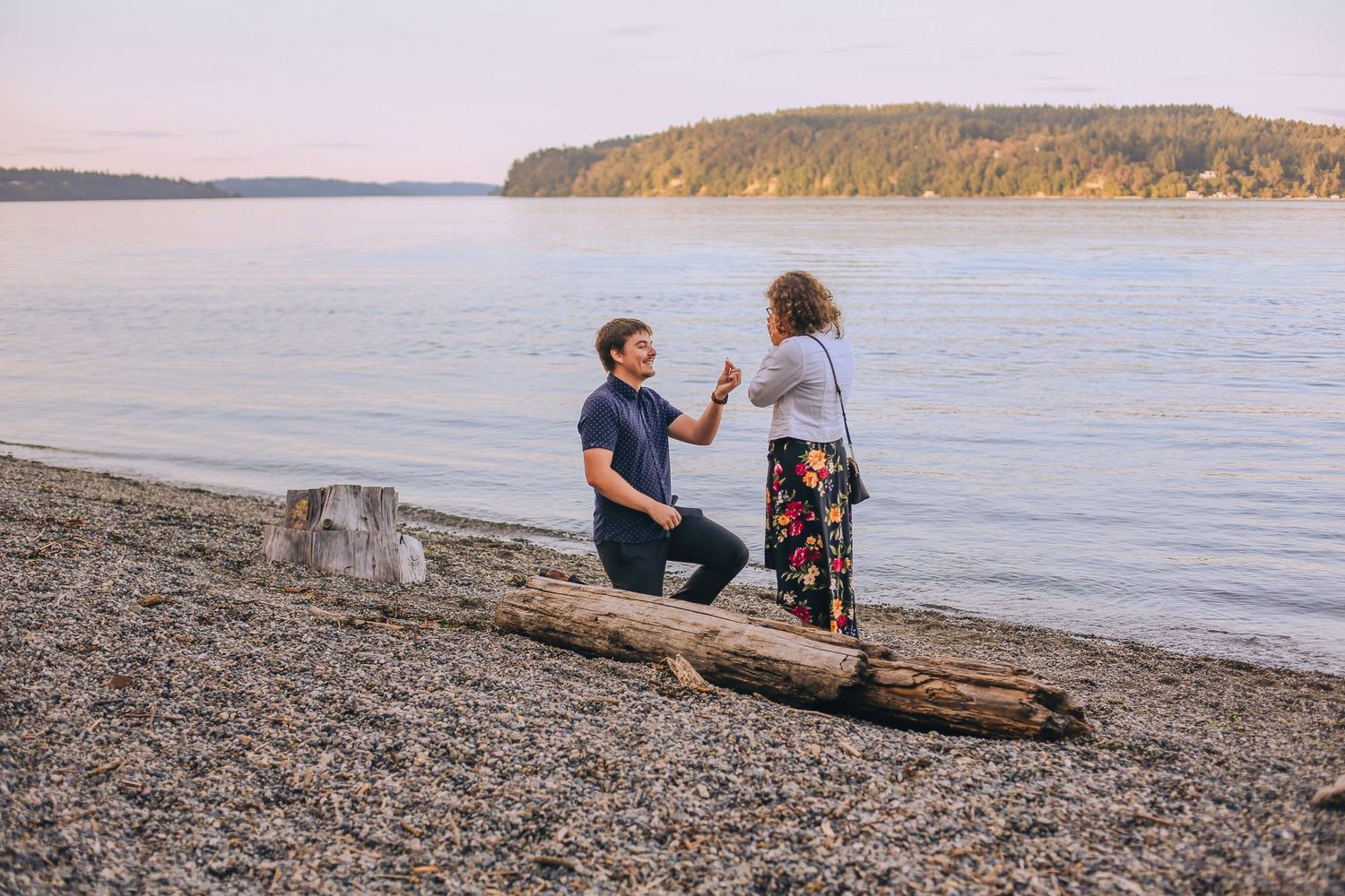 Couple sitting on driftwood at sunset on a pebble beach with mountains in the distance. Spokane Liberty Lake Photographer Family Photographe
