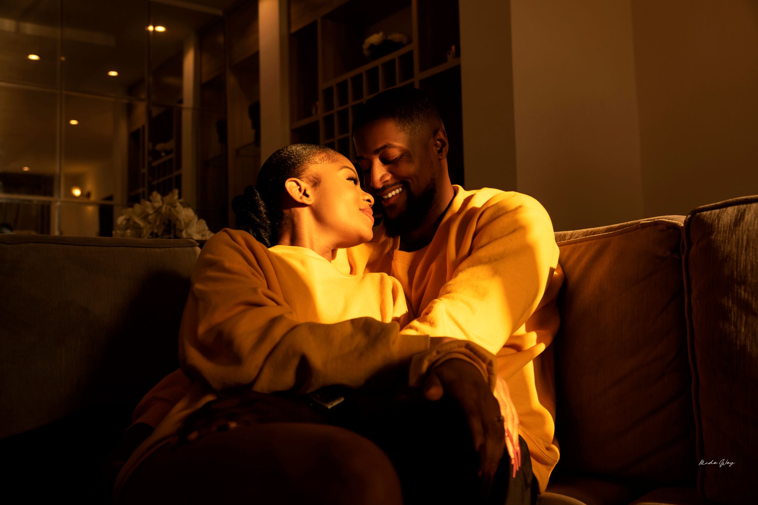 Couple shares intimate moment on brown leather couch in warm, dim lighting.
