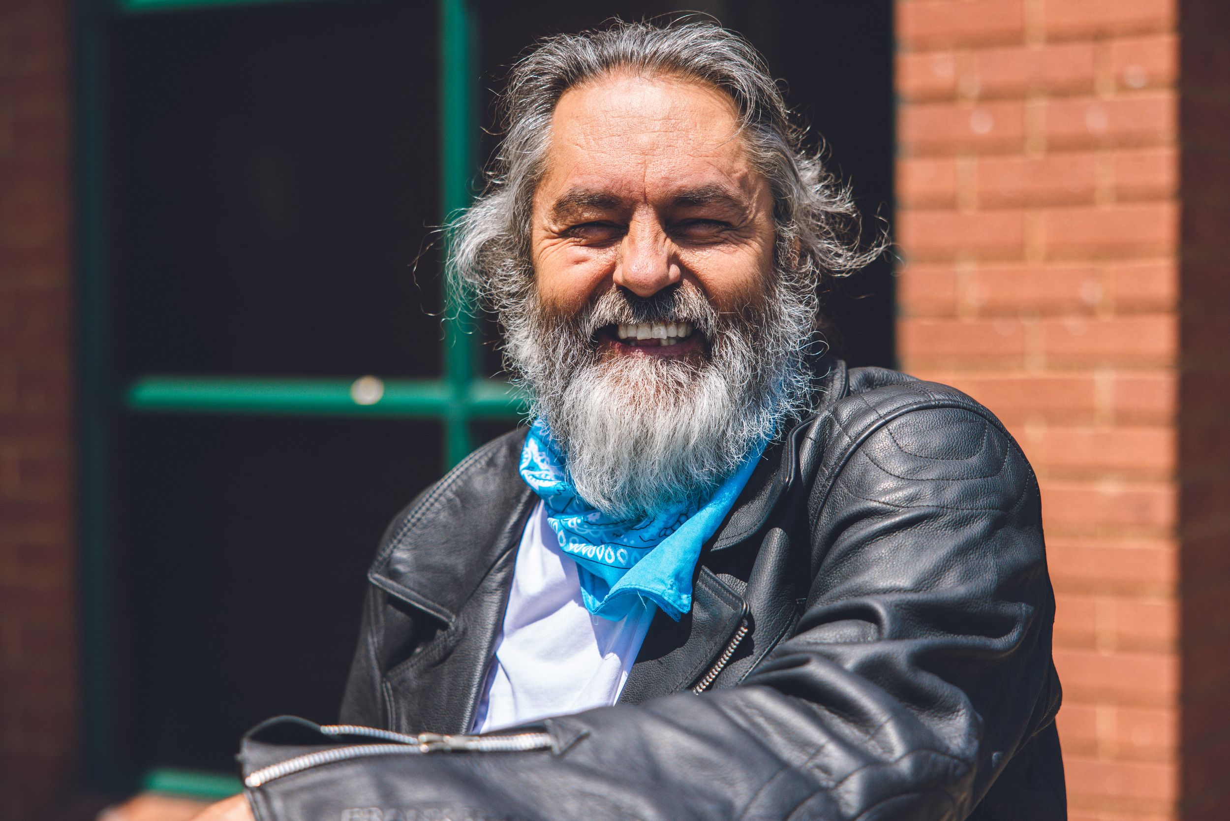 Older man with grey beard wearing leather jacket and blue scarf smiles at camera.