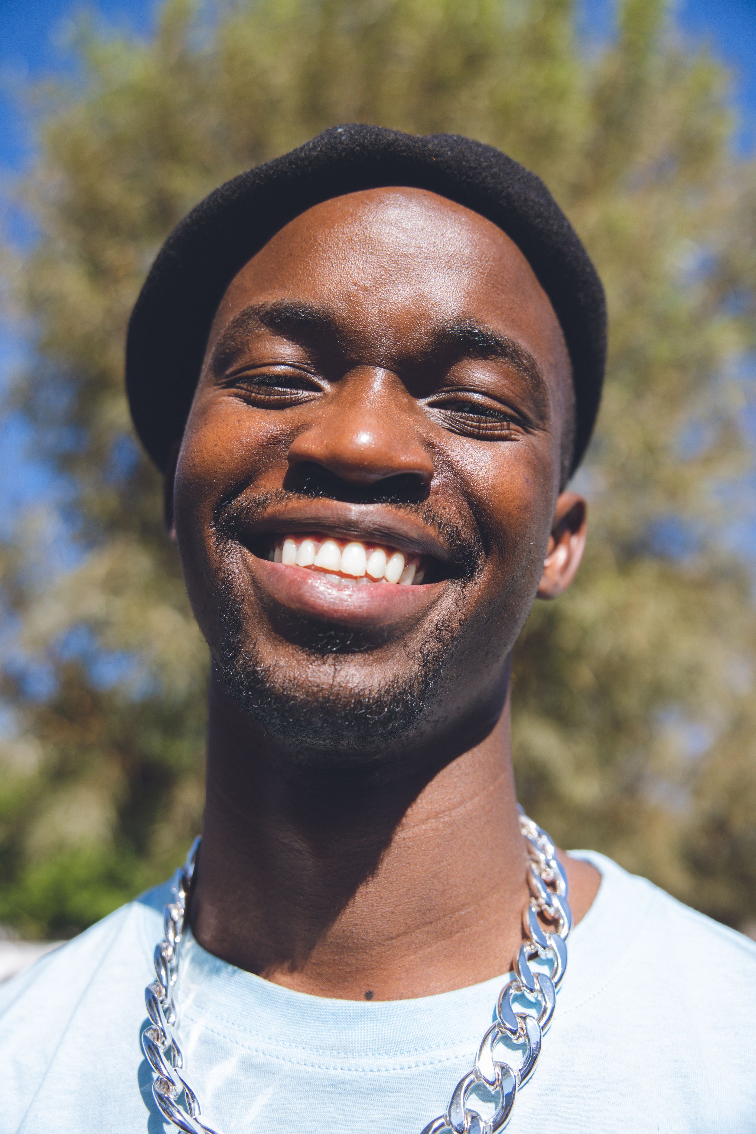 Young man wearing beret and chain necklace smiles brightly in outdoor sunlit setting.