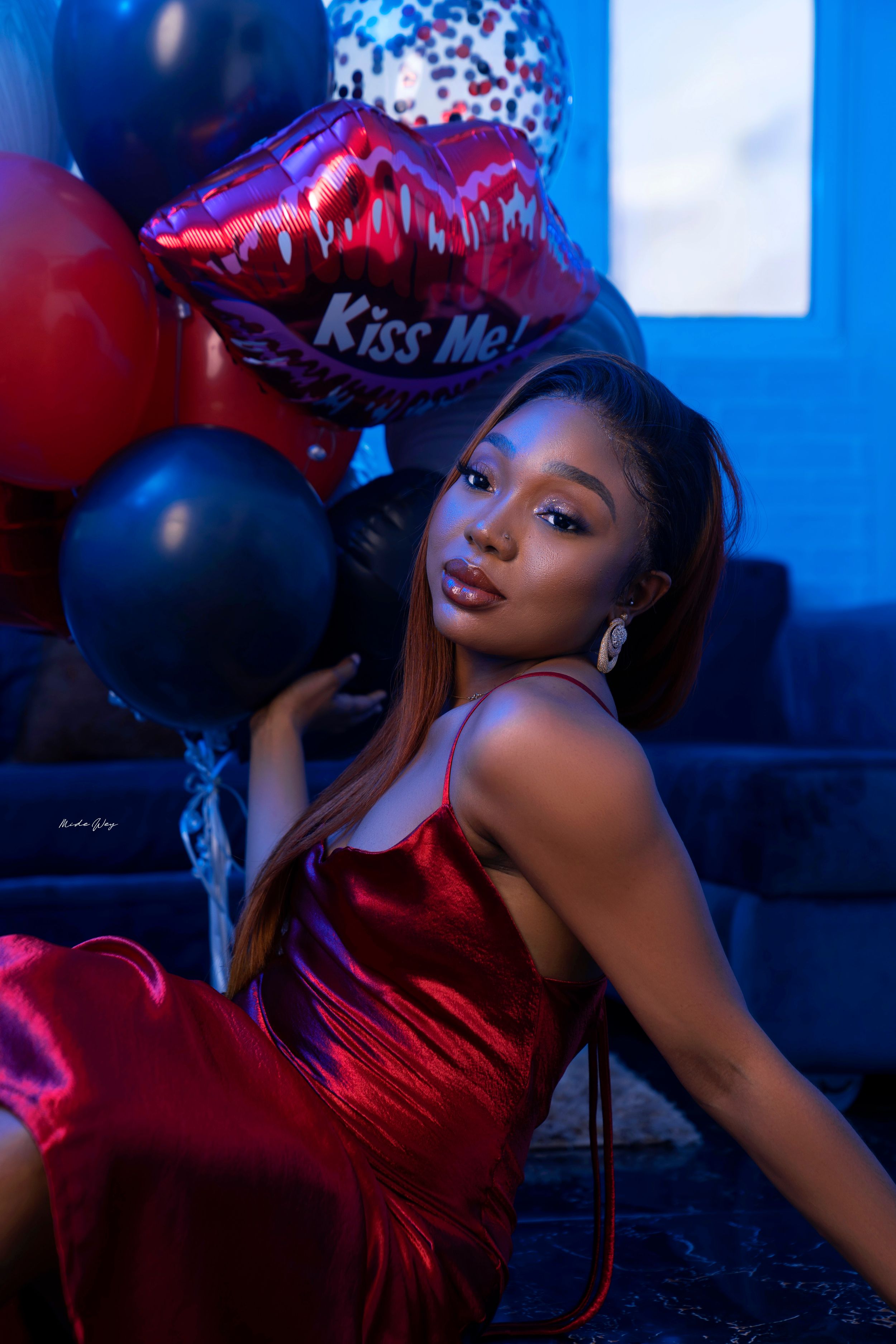 Woman in red satin dress poses with Valentine-themed balloons in moody blue lighting.