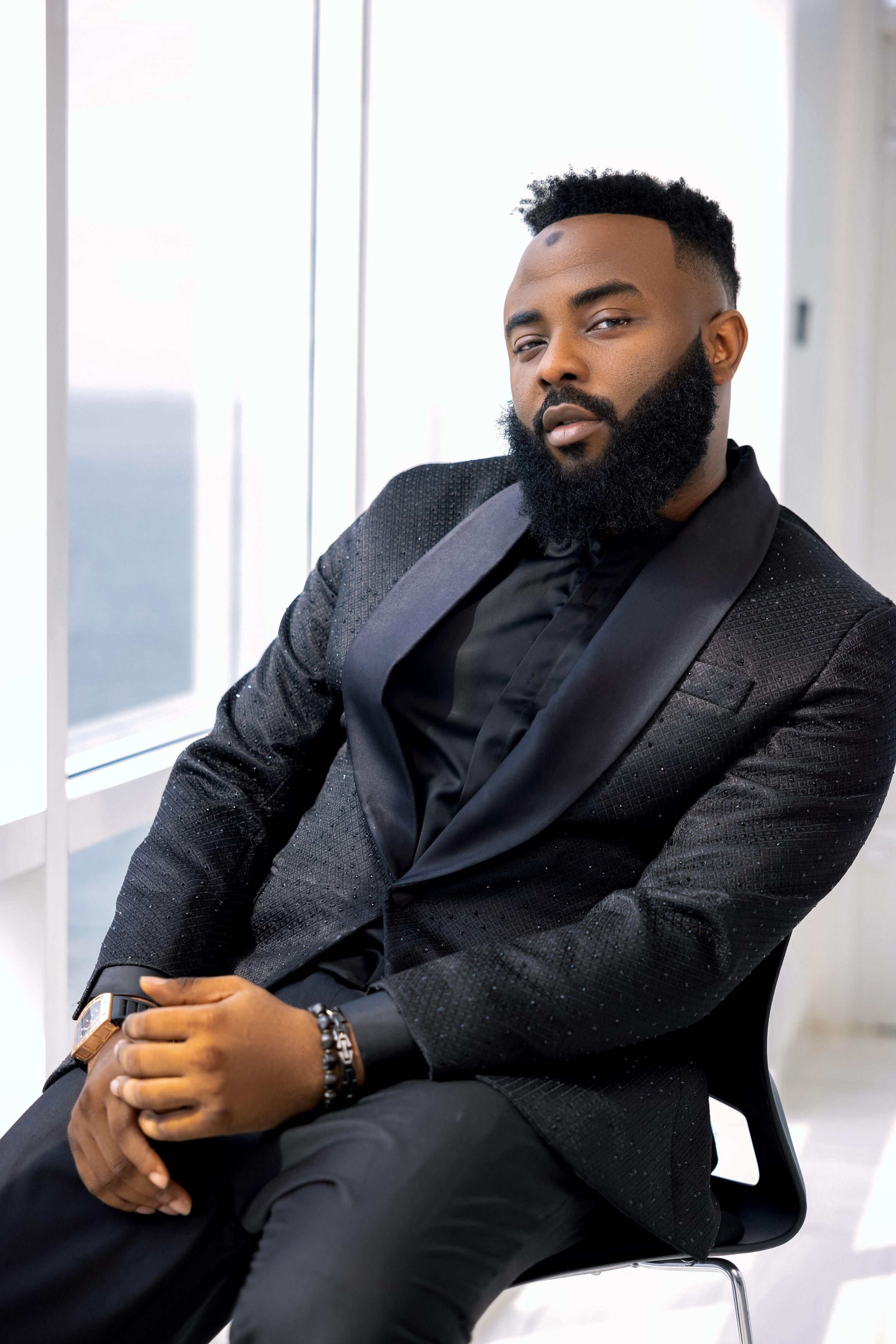 Professional portrait of well-dressed person in dark suit sitting thoughtfully by window.