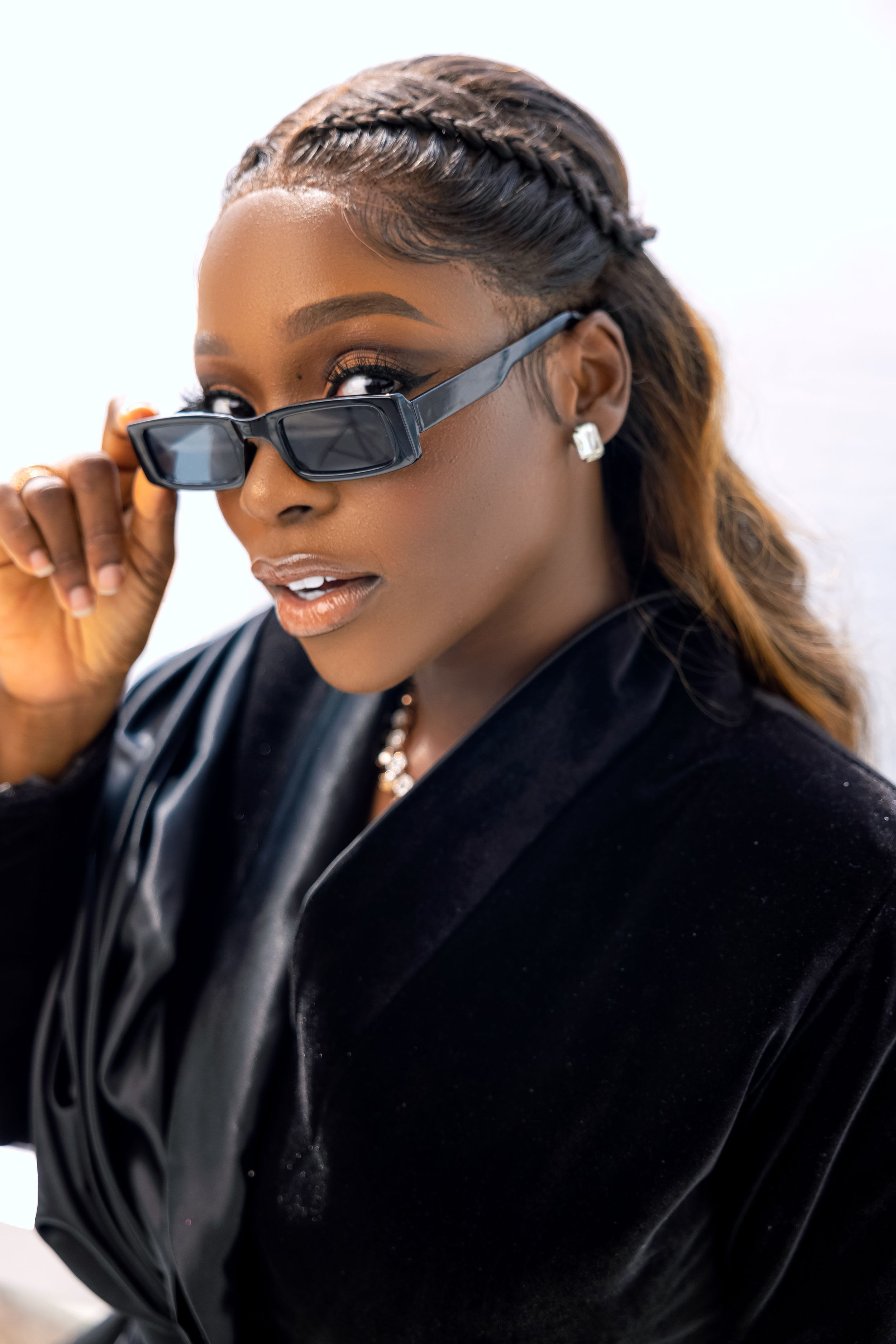 Stylish portrait featuring braided hair, dark sunglasses and black blazer in natural lighting.