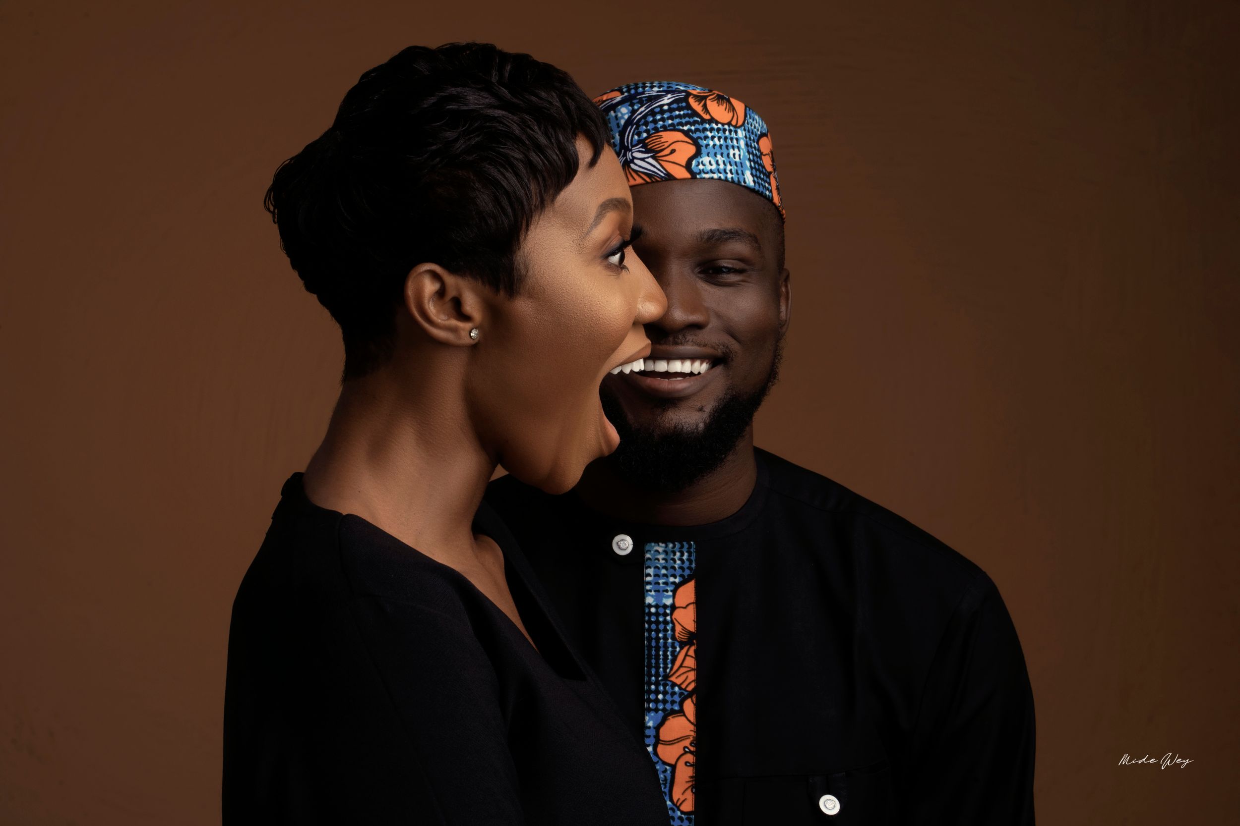 A couple sharing a candid laughing moment against a brown studio backdrop.