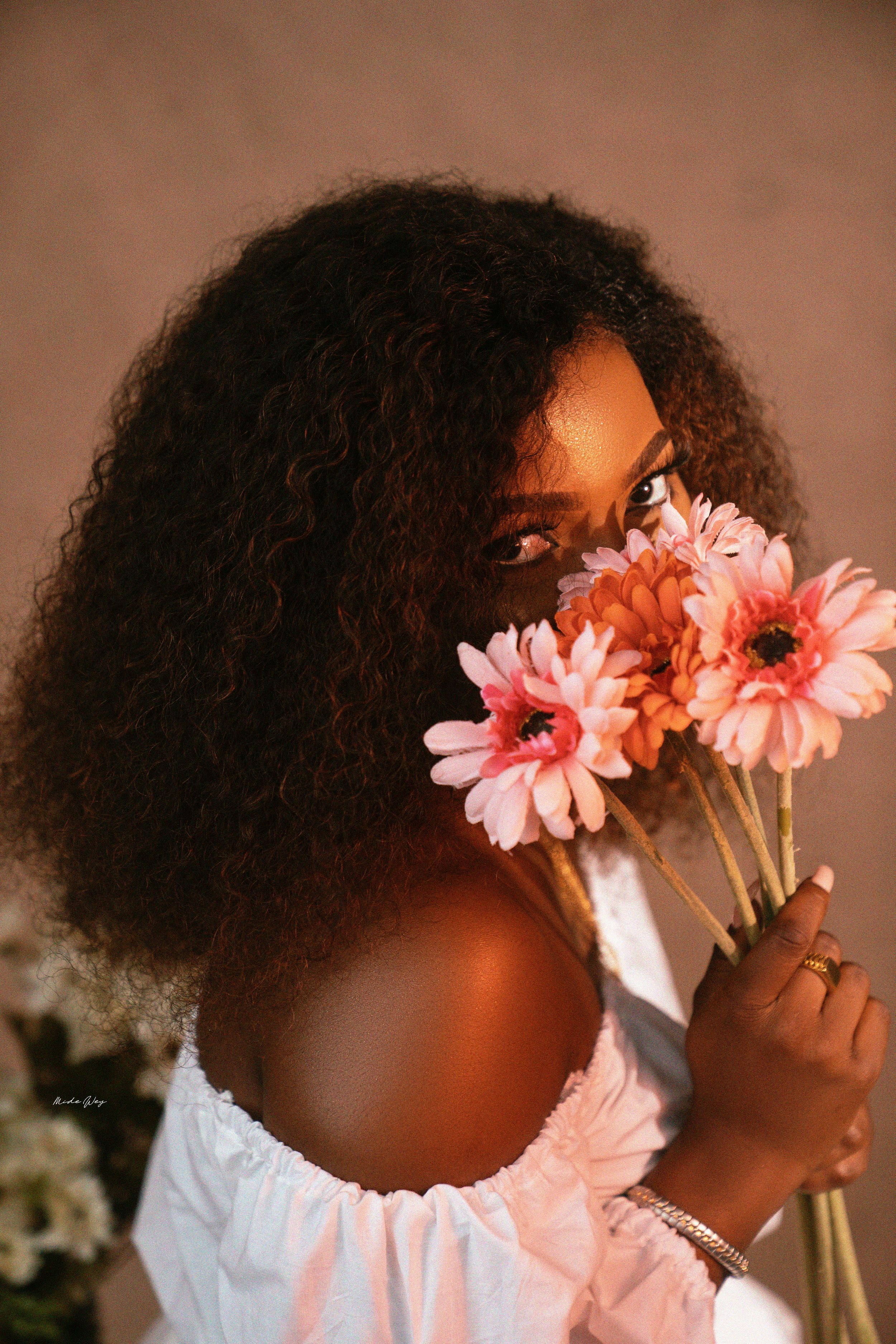 Artistic portrait with natural curls holding pink gerbera daisies against warm background.