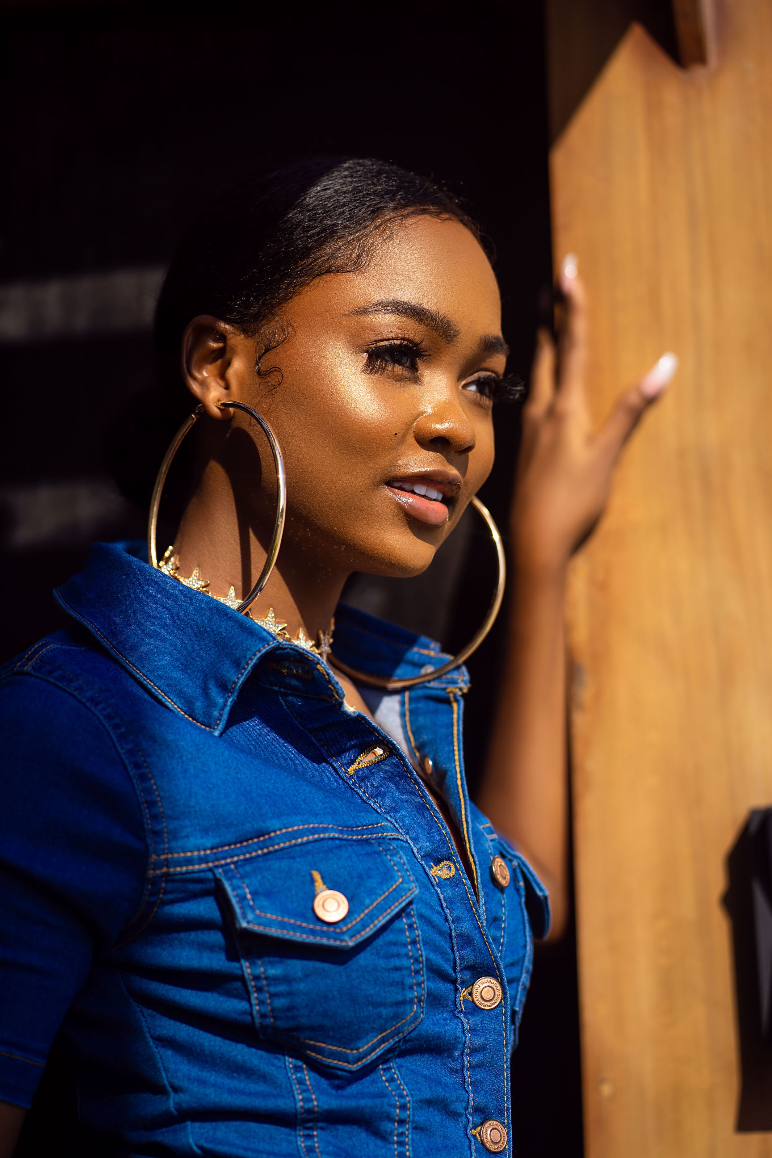 Woman in blue denim jacket and gold hoop earrings poses against wooden wall in dramatic lighting.