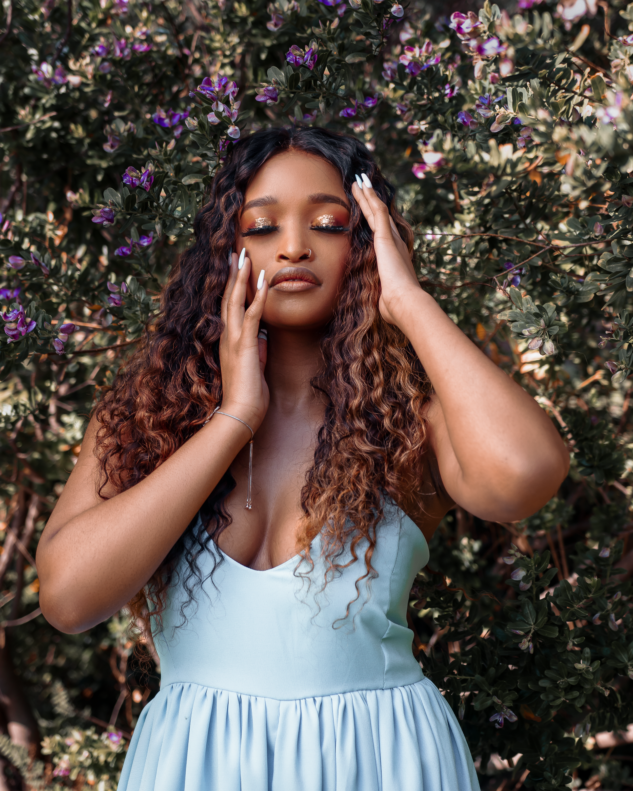 Person in light blue dress with curly hair poses against flowering bush background.