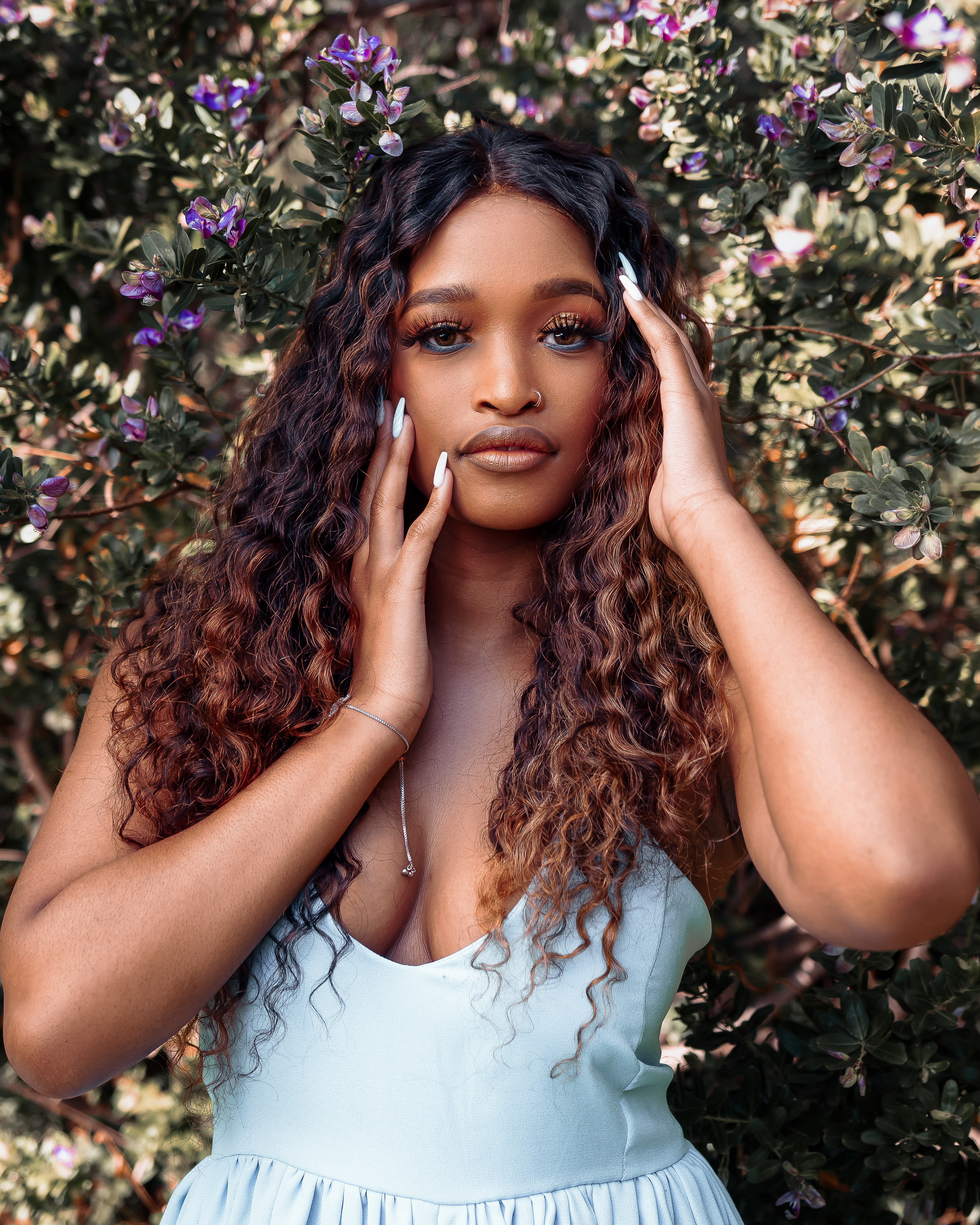 Portrait against flowering bushes showing soft makeup and flowing curls.