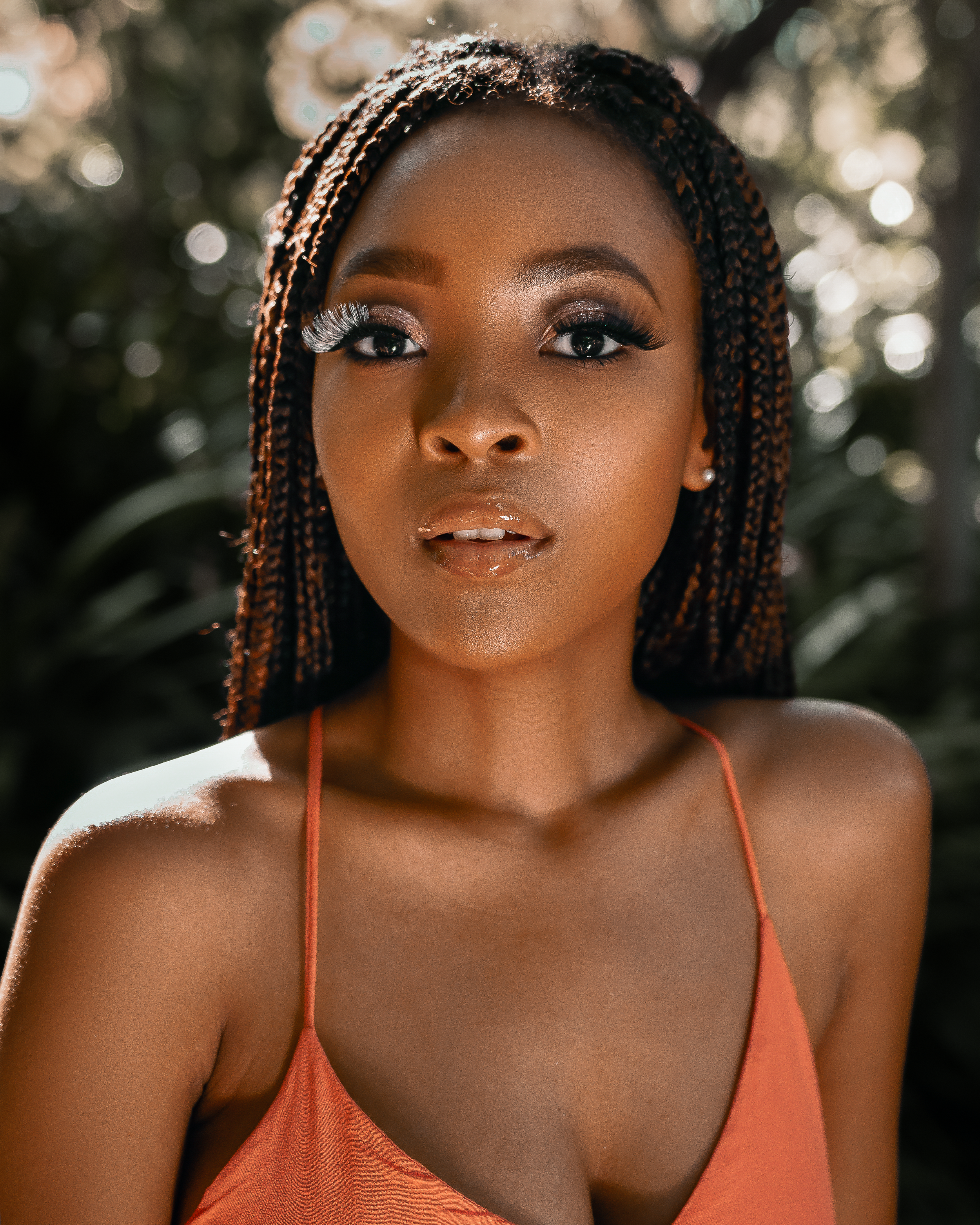 Portrait in orange tank top with braided hair against blurred natural backdrop in golden sunlight.