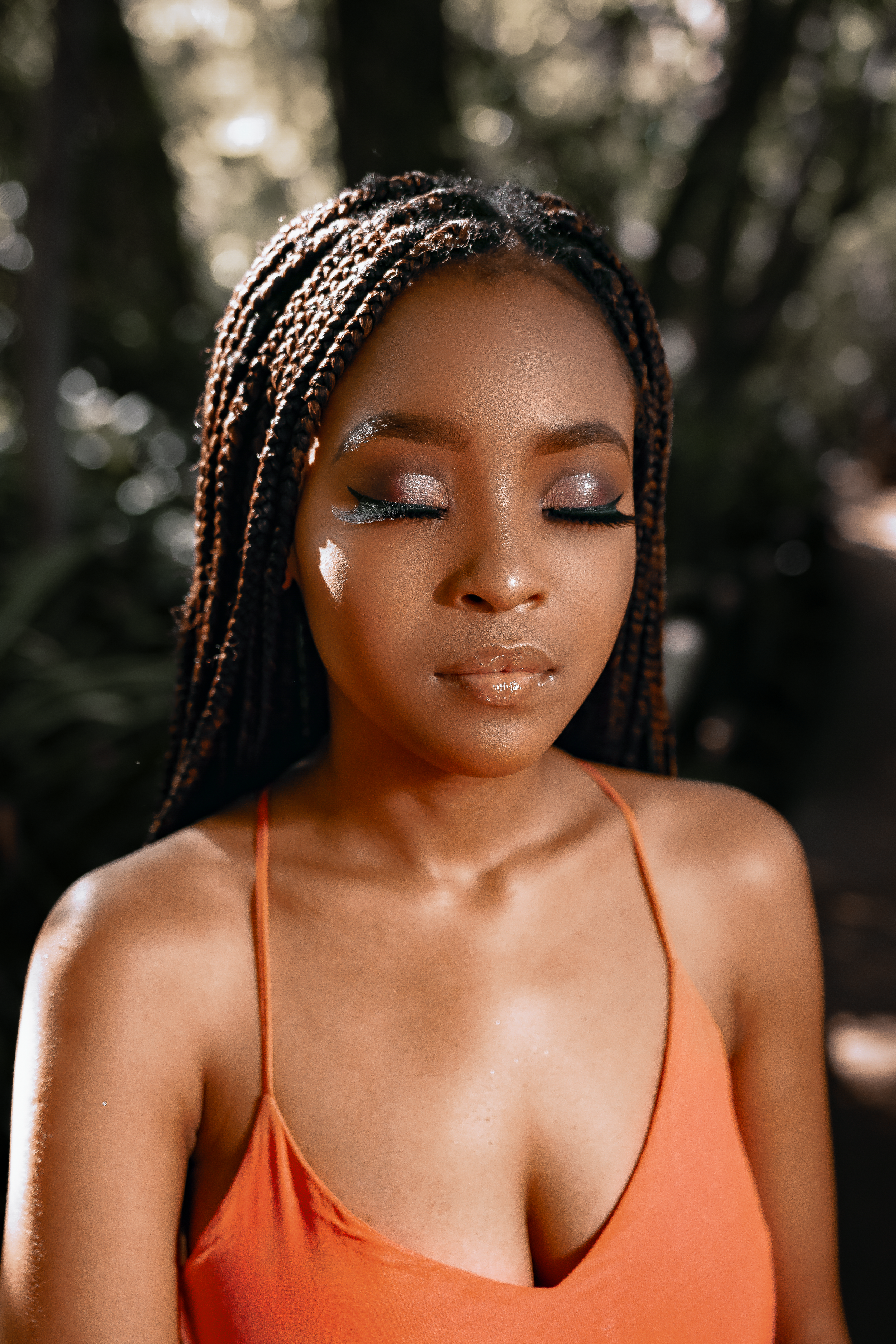Artistic makeup portrait with glitter details and closed eyes in orange top against bokeh background.