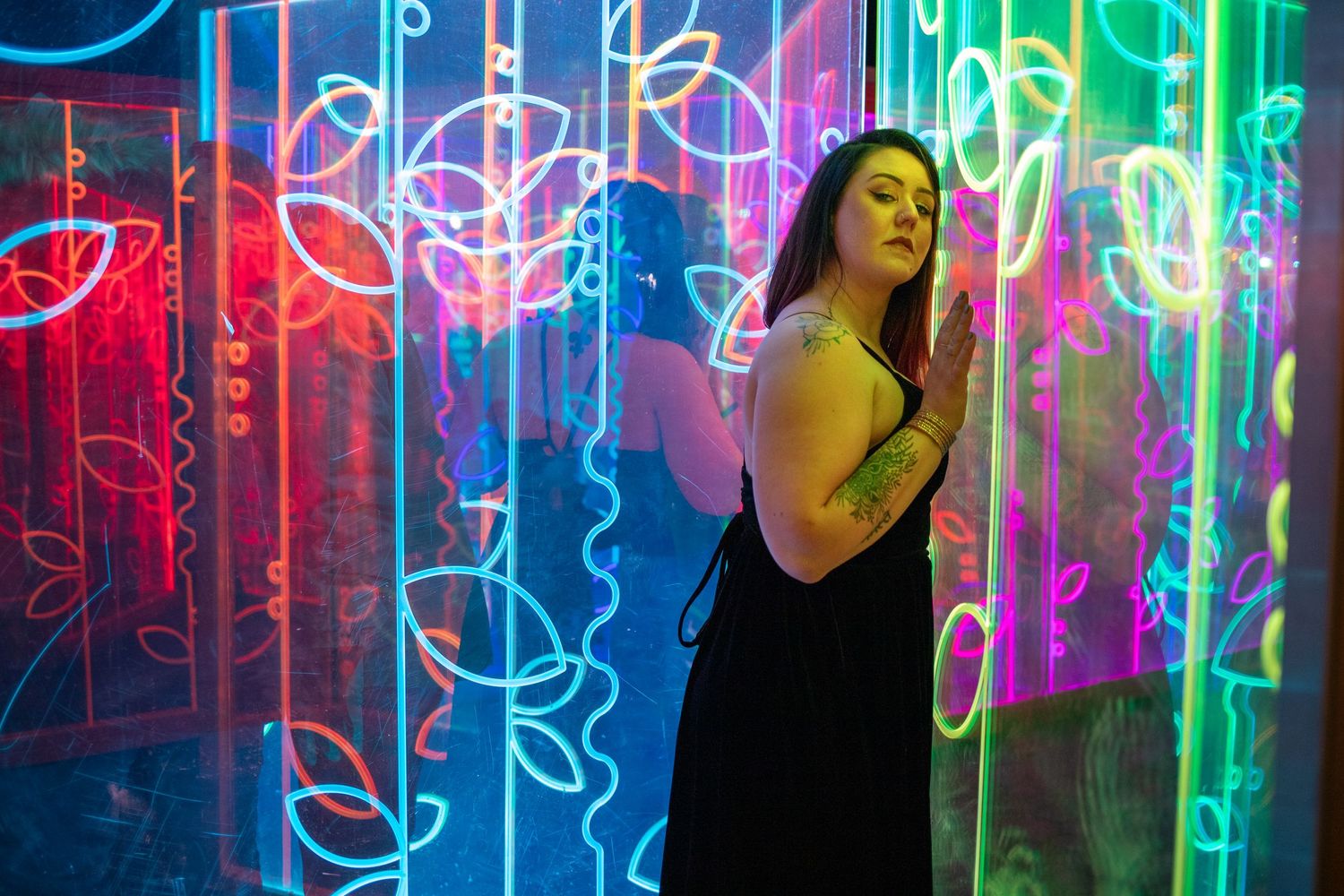 Woman in black dress poses thoughtfully against vibrant colorful neon light installation backdrop.