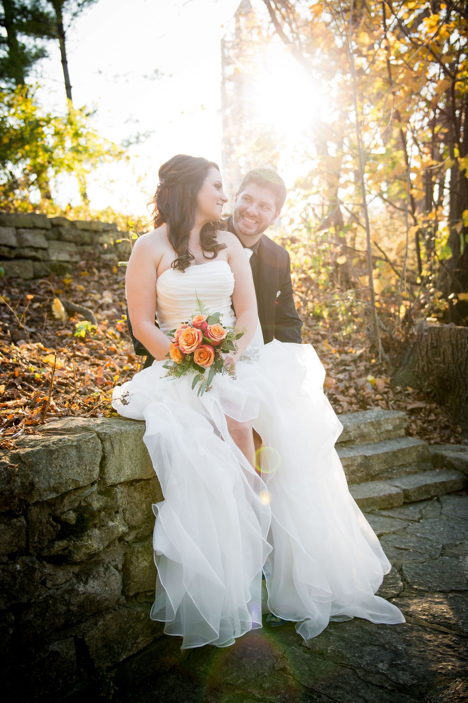 Bride and Groom sitting on a stone ledge with sunlight shining through colored leaves during fall in Naperville, IL.
