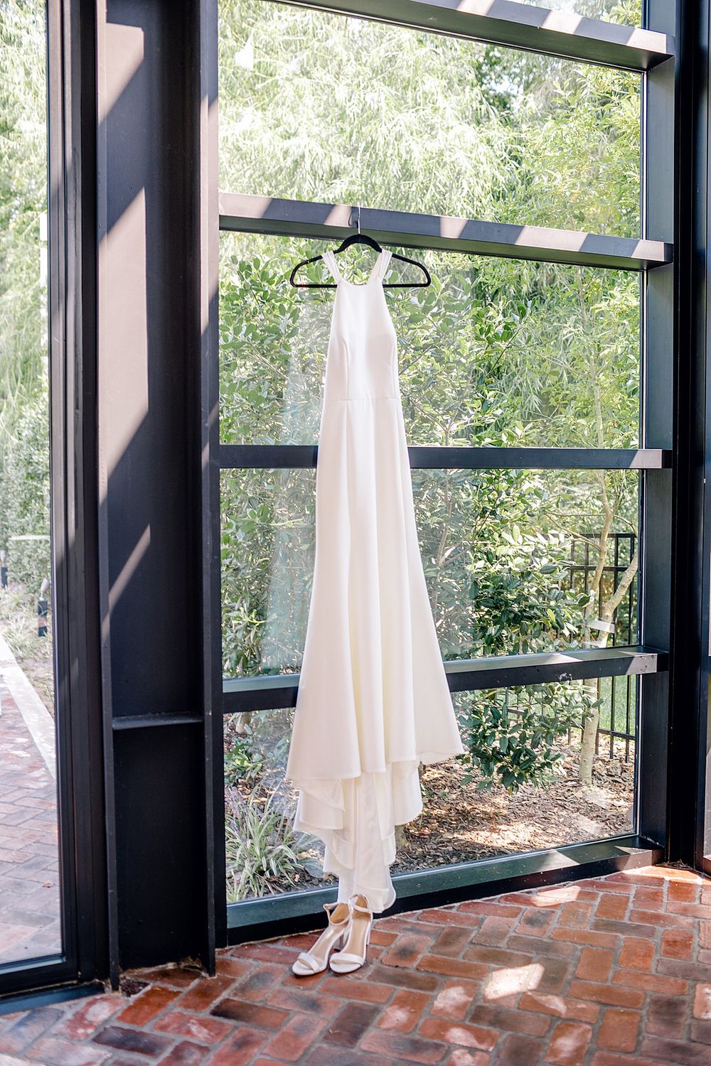 Elegant white wedding gown hangs in window with natural light streaming through.