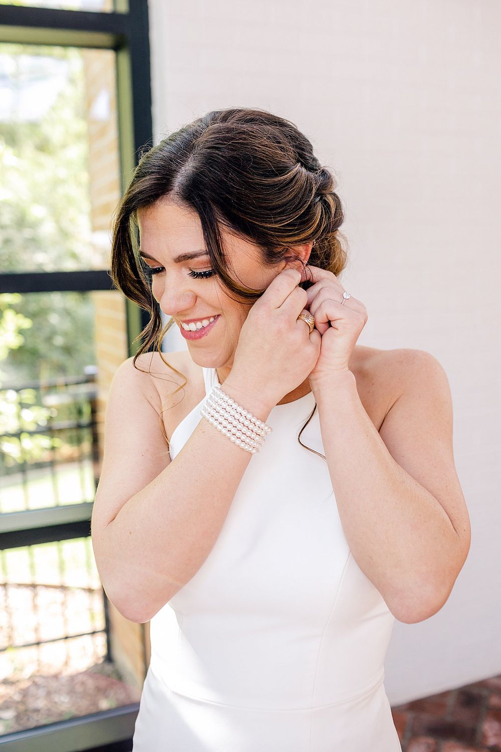 An elegant bride wearing a pearl bracelet and white halter dress smiles while putting on earrings.