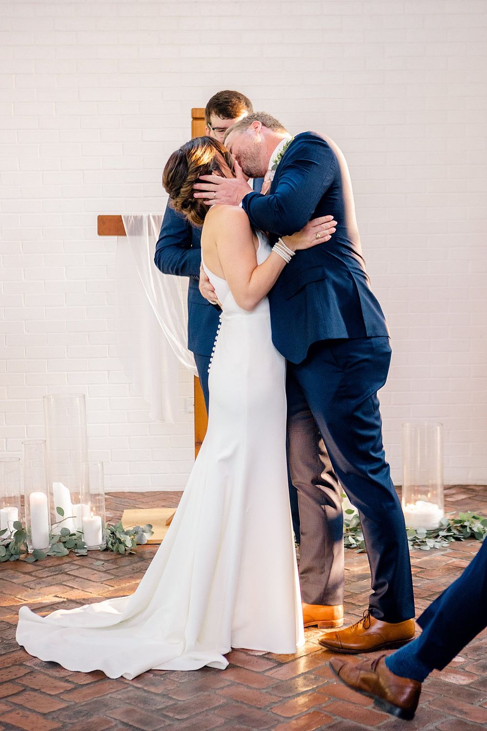 Romantic first kiss moment between newlyweds in front of a white cross with glowing candles.