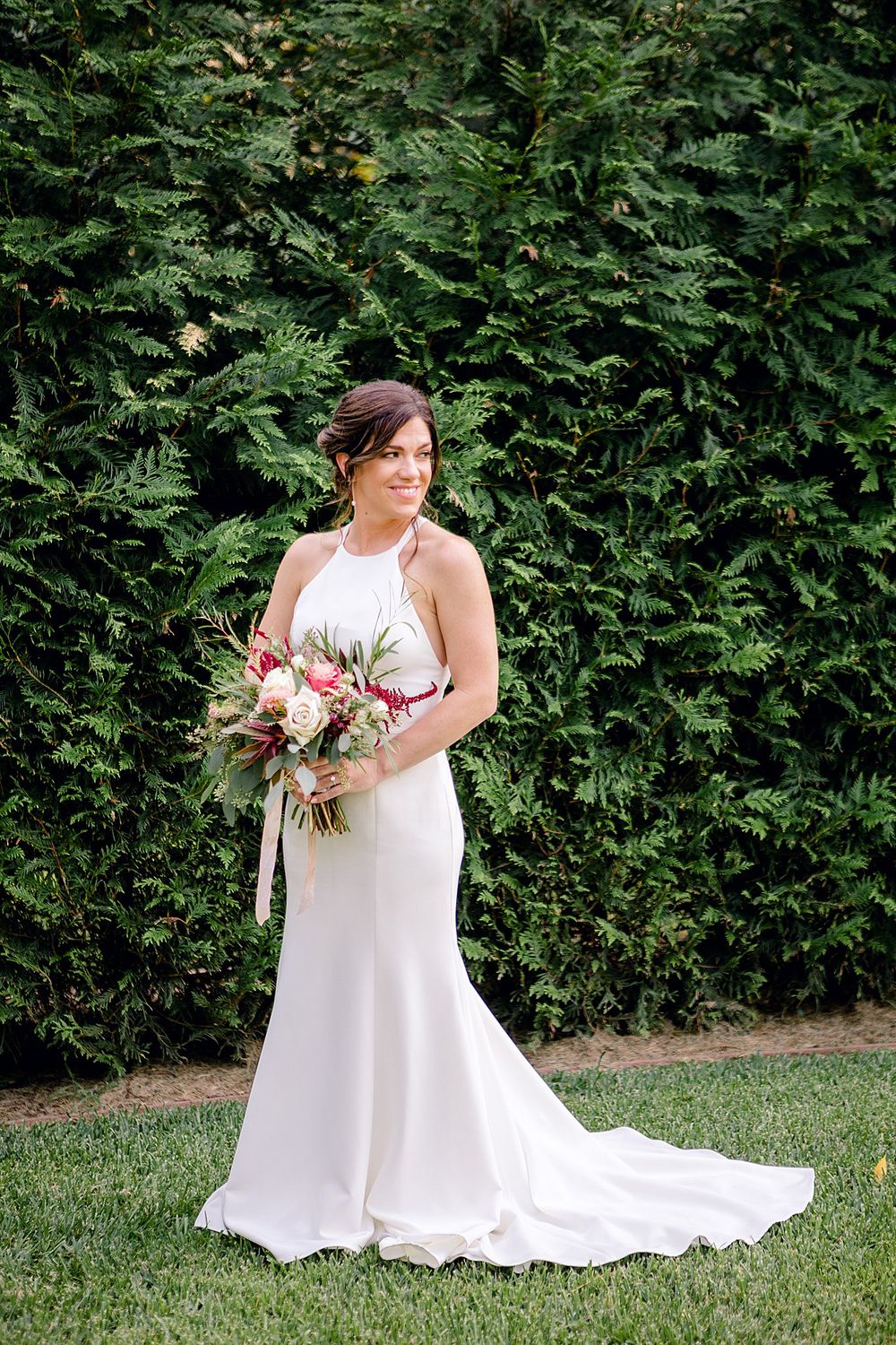 Elegant bride in white halter dress poses with bouquet against lush green hedge backdrop.