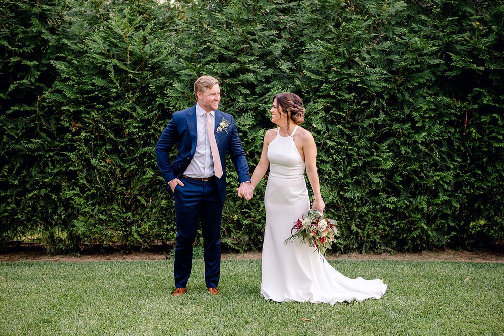 Wedding couple holding hands while standing on lawn in front of evergreen hedge.