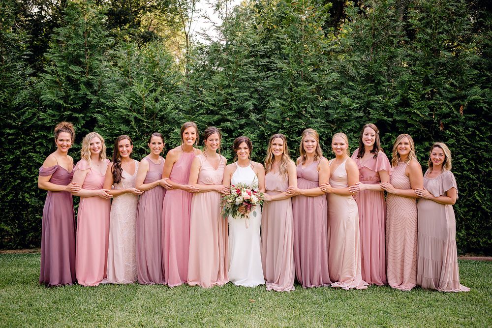 Bridal party wearing various shades of pink and blush dresses lined up outdoors.